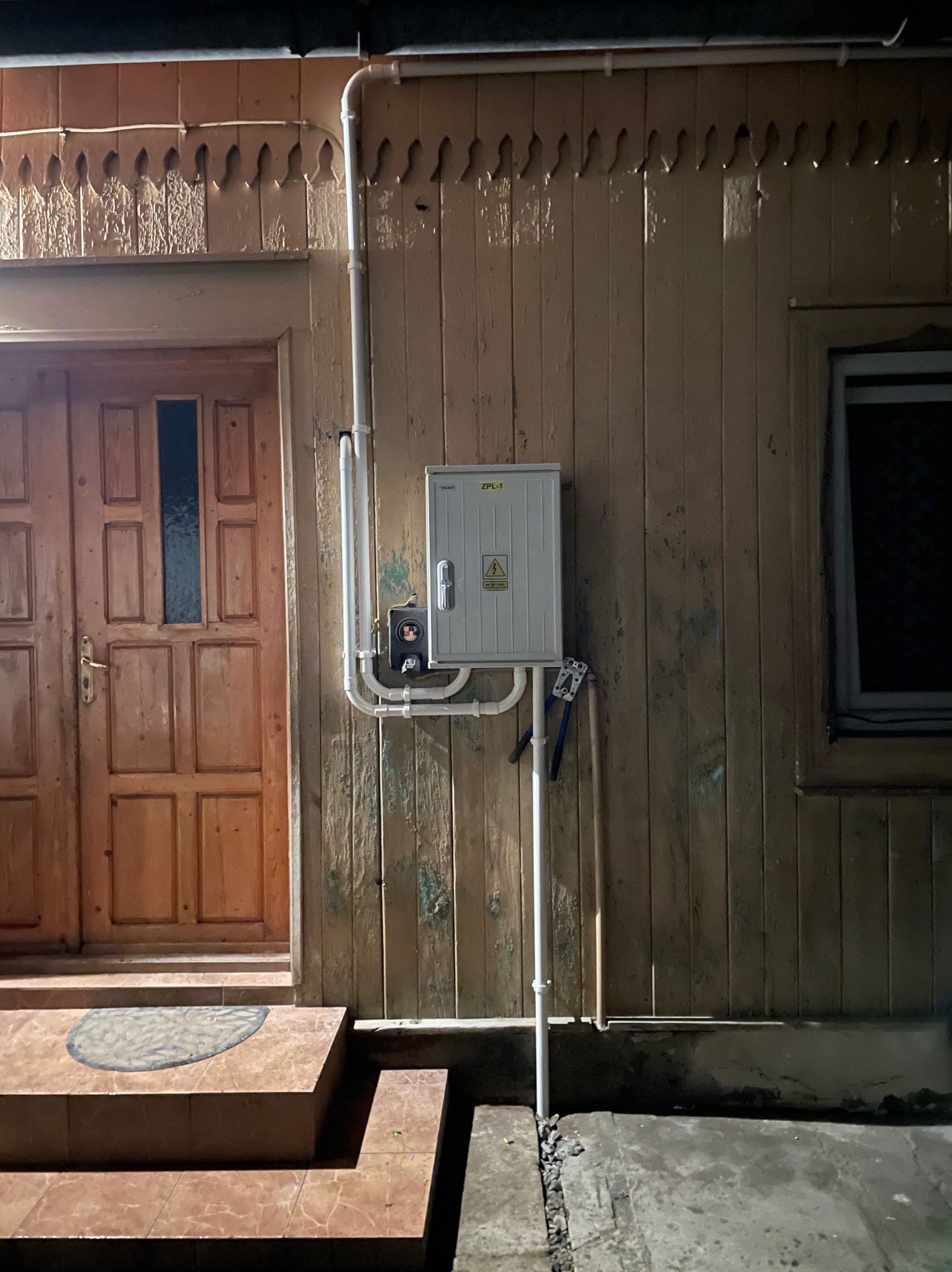 Zewnętrzna skrzynka elektryczna ZPL-1 z ostrzeżeniem o napięciu, zamontowana na drewnianej elewacji domu z widocznymi rurami instalacyjnymi i narzędziami elektrycznymi obok wejścia.