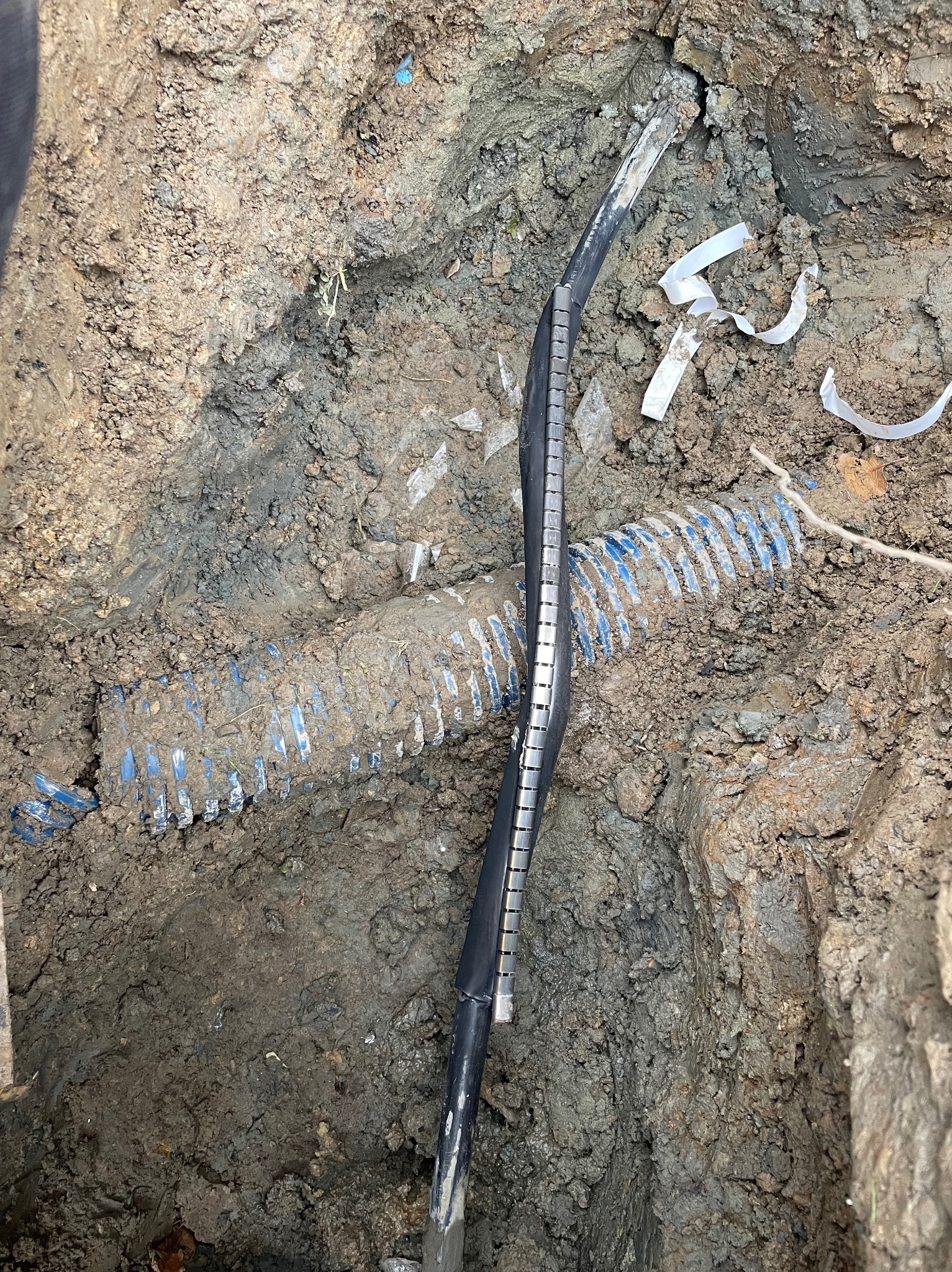 Kabel elektryczny w ziemi, obok niebieskiej rury karbowanej, w wykopie z gliniastej ziemi, z fragmentami taśmy izolacyjnej.