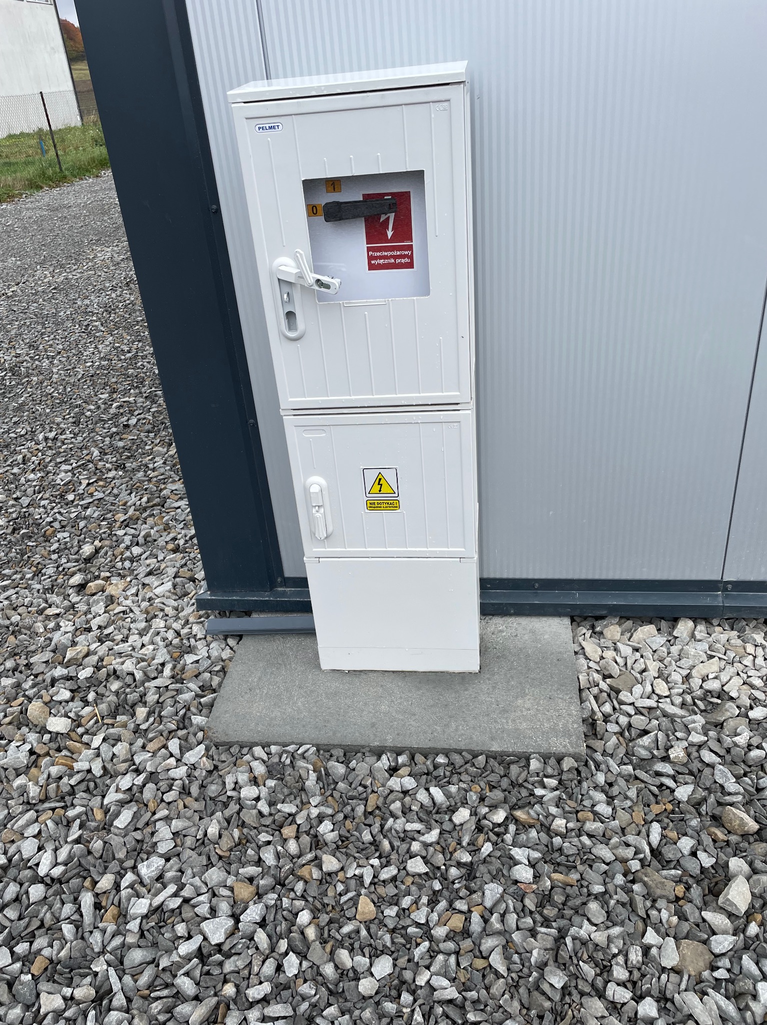 Biała skrzynka elektryczna z ostrzeżeniem o wysokim napięciu, zamontowana na szarej, metalowej ścianie budynku, na betonowej wylewce otoczonej żwirem.