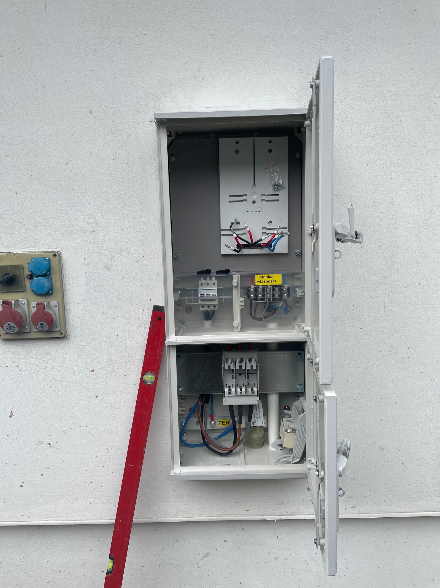 Otwarta skrzynka elektryczna na białej ścianie, widoczne zabezpieczenia, przewody, listwa zaciskowa, poziomica oparta o ścianę obok skrzynki. Z boku gniazda siłowe.