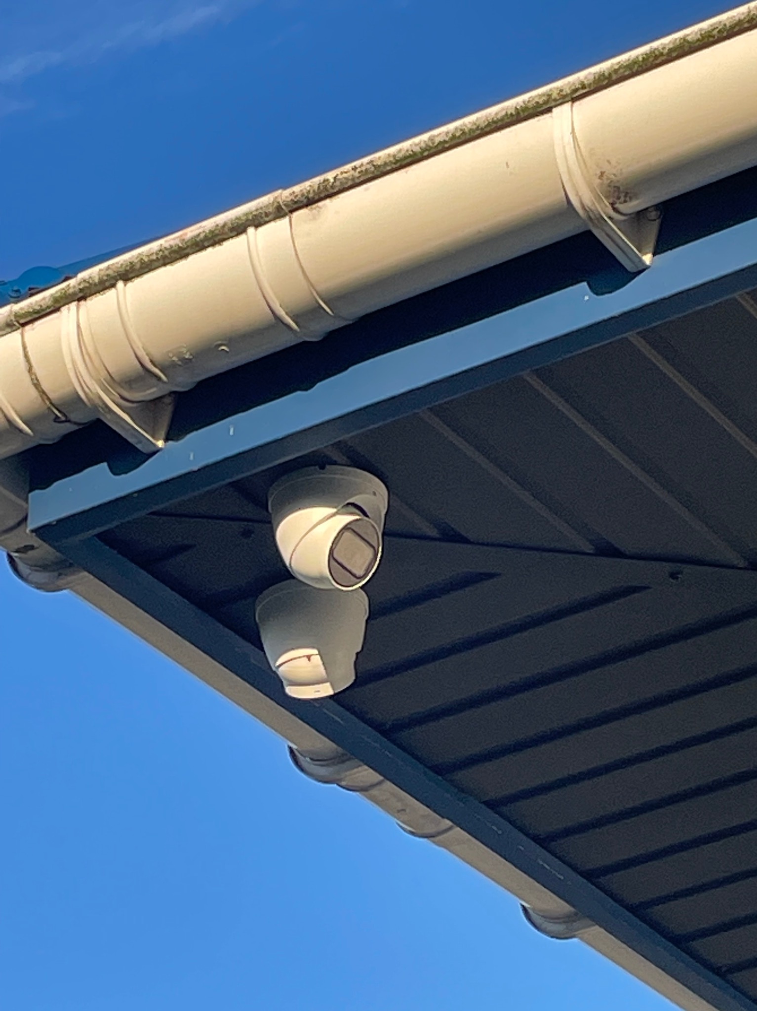 Kamera monitoringu zamontowana pod okapem budynku, widok z dołu na tle błękitnego nieba.