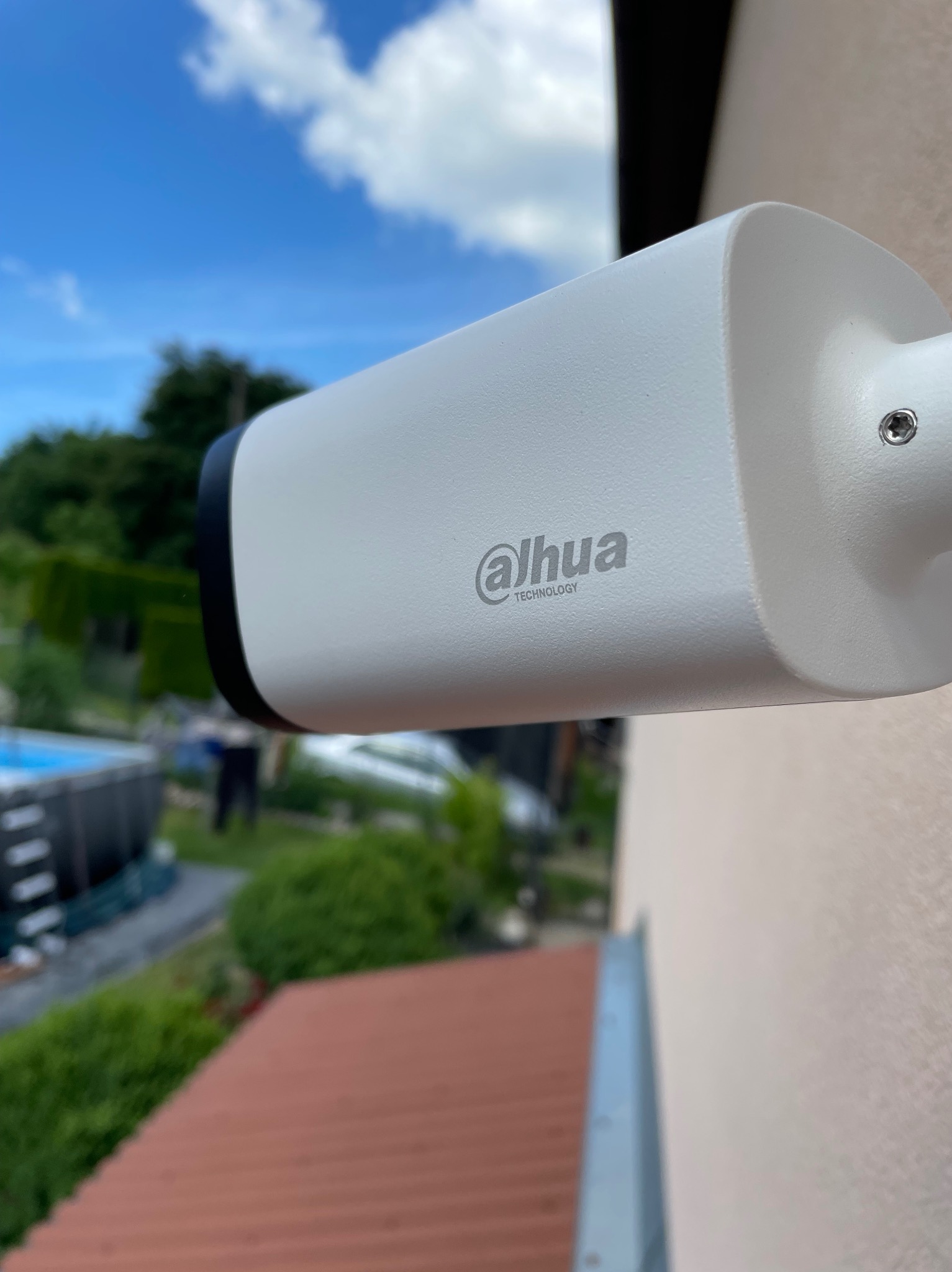 Zewnętrzna kamera monitoringu Dahua zamontowana na elewacji domu, widok z boku, w tle błękitne niebo i zieleń ogrodu z basenem.