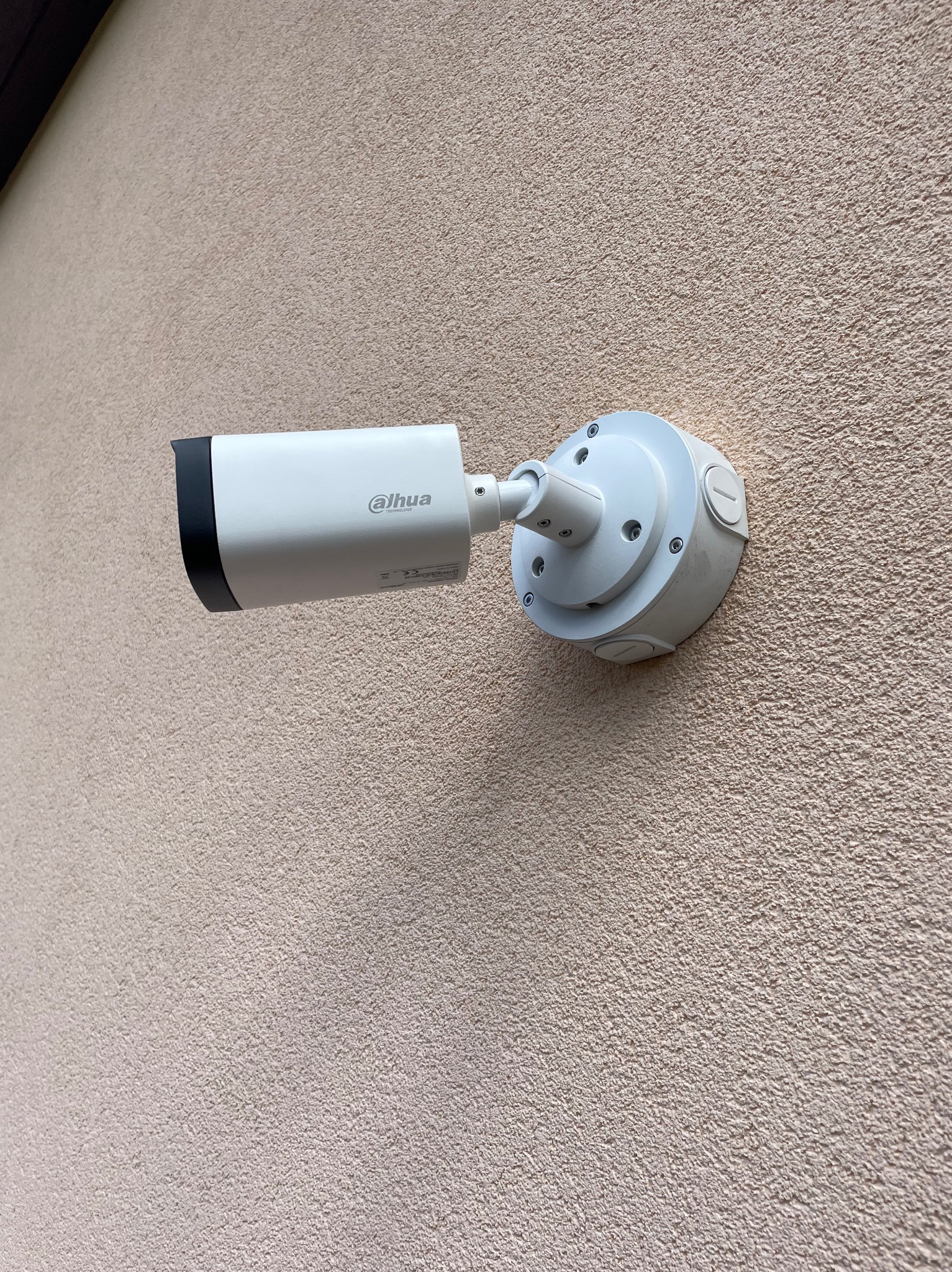 Biała kamera monitoringu Dahua zamontowana na beżowej, chropowatej ścianie budynku, widoczne śruby mocujące i logo producenta.