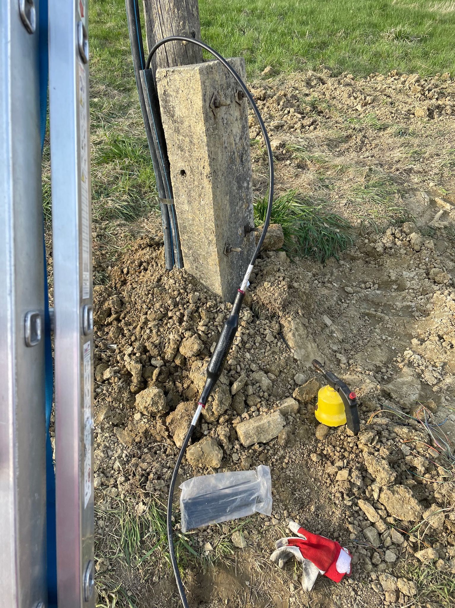 Słup betonowy z przymocowanymi kablami, widoczne narzędzia, złączka kablowa, rękawice robocze i drabina w tle, teren ziemisty.