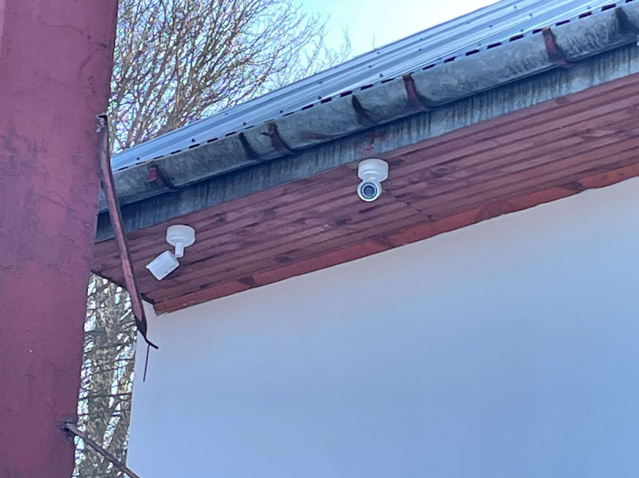 Dwie białe kamery monitoringu zamontowane pod okapem budynku z drewnianą podbitką; jedna kamera skierowana w dół, druga na bok, widoczny komin i drzewo bez liści w tle.