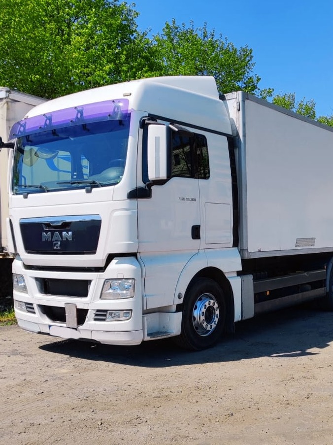 Biała ciężarówka MAN TGS 26.360 z fioletową osłoną przeciwsłoneczną zaparkowana w Łodzi na żwirowym placu, widoczna z przodu pod lekkim kątem.
