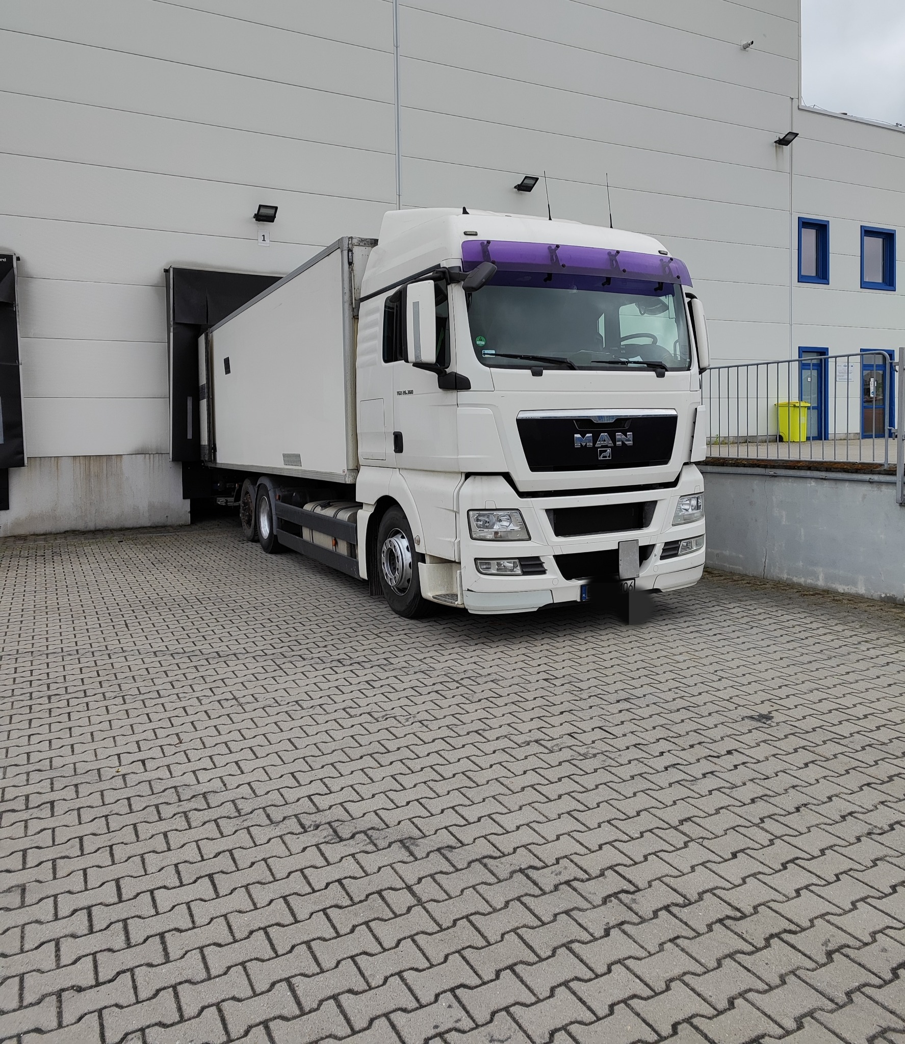 Biała ciężarówka MAN z fioletową blendą zaparkowana przy rampie magazynowej z szarej kostki brukowej.
