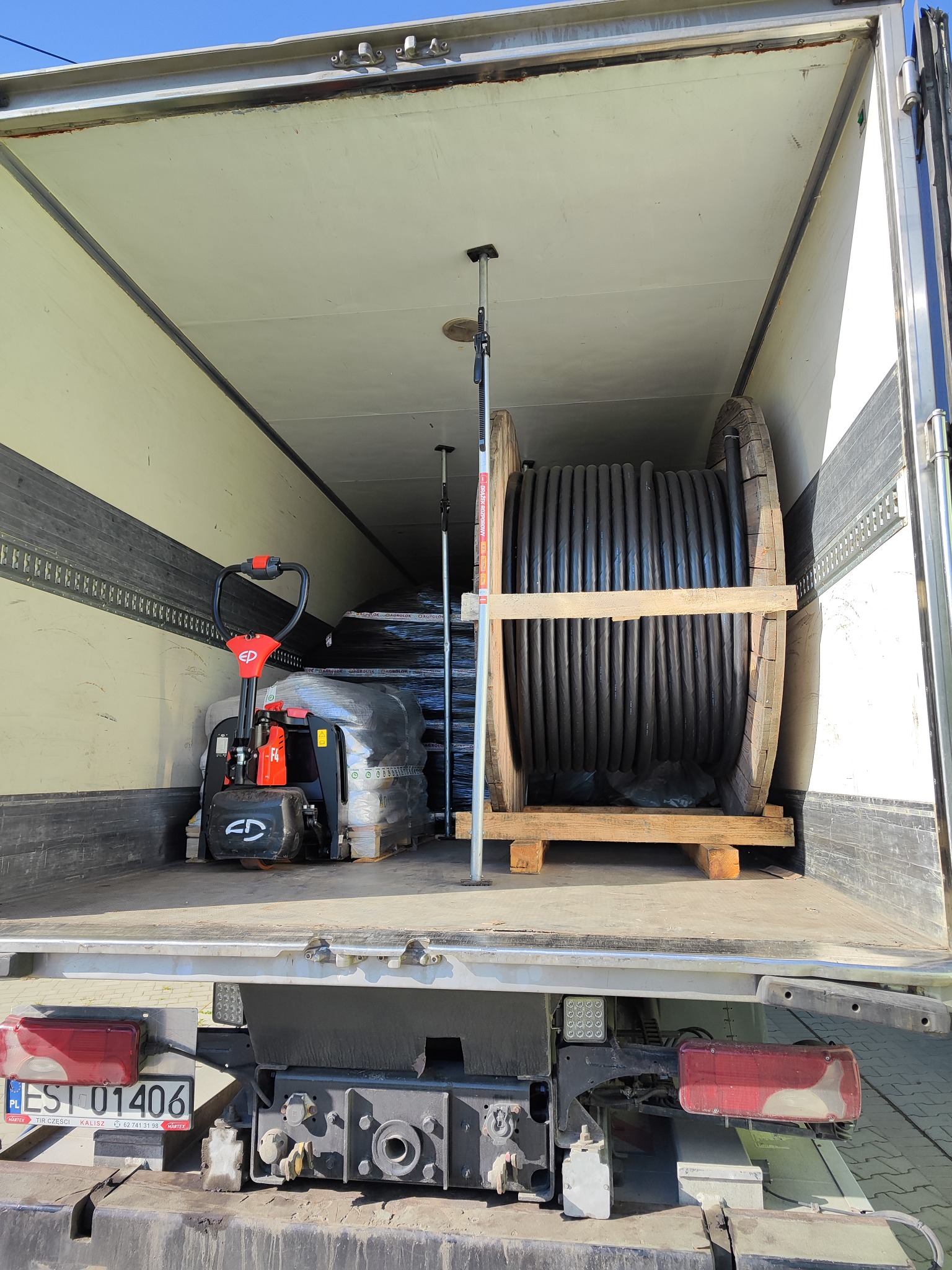 Wnętrze naczepy ciężarówki z ładunkiem: duża szpula z czarnym kablem na drewnianej palecie i paleciak z owiniętym towarem, zabezpieczone rozpórkami.