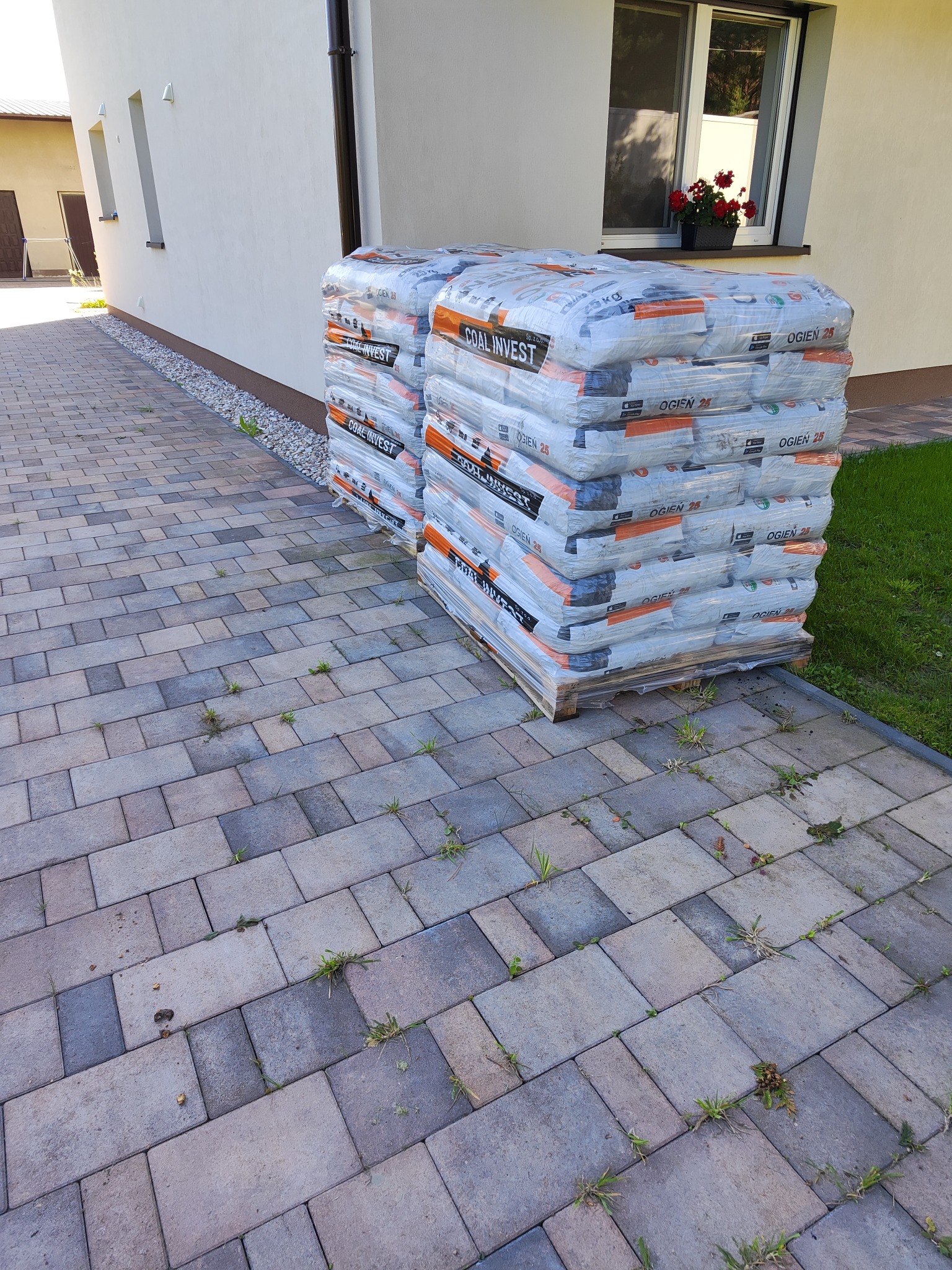 Dwa palety z workami opału 'OGIEŃ 25' w folii stoją na kostce brukowej przed jasnym budynkiem z oknem, w którym widać kwiaty w doniczce.