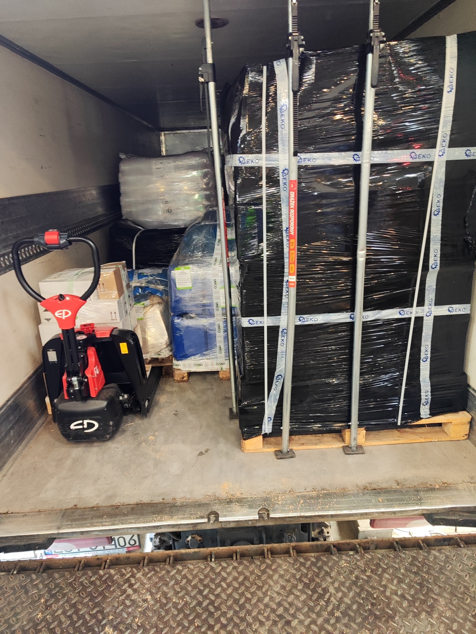 Wnętrze przestrzeni ładunkowej samochodu ciężarowego z załadowanym towarem na paletach, zabezpieczonym pasami transportowymi i drążkami rozporowymi, widoczny wózek paletowy elektryczny.