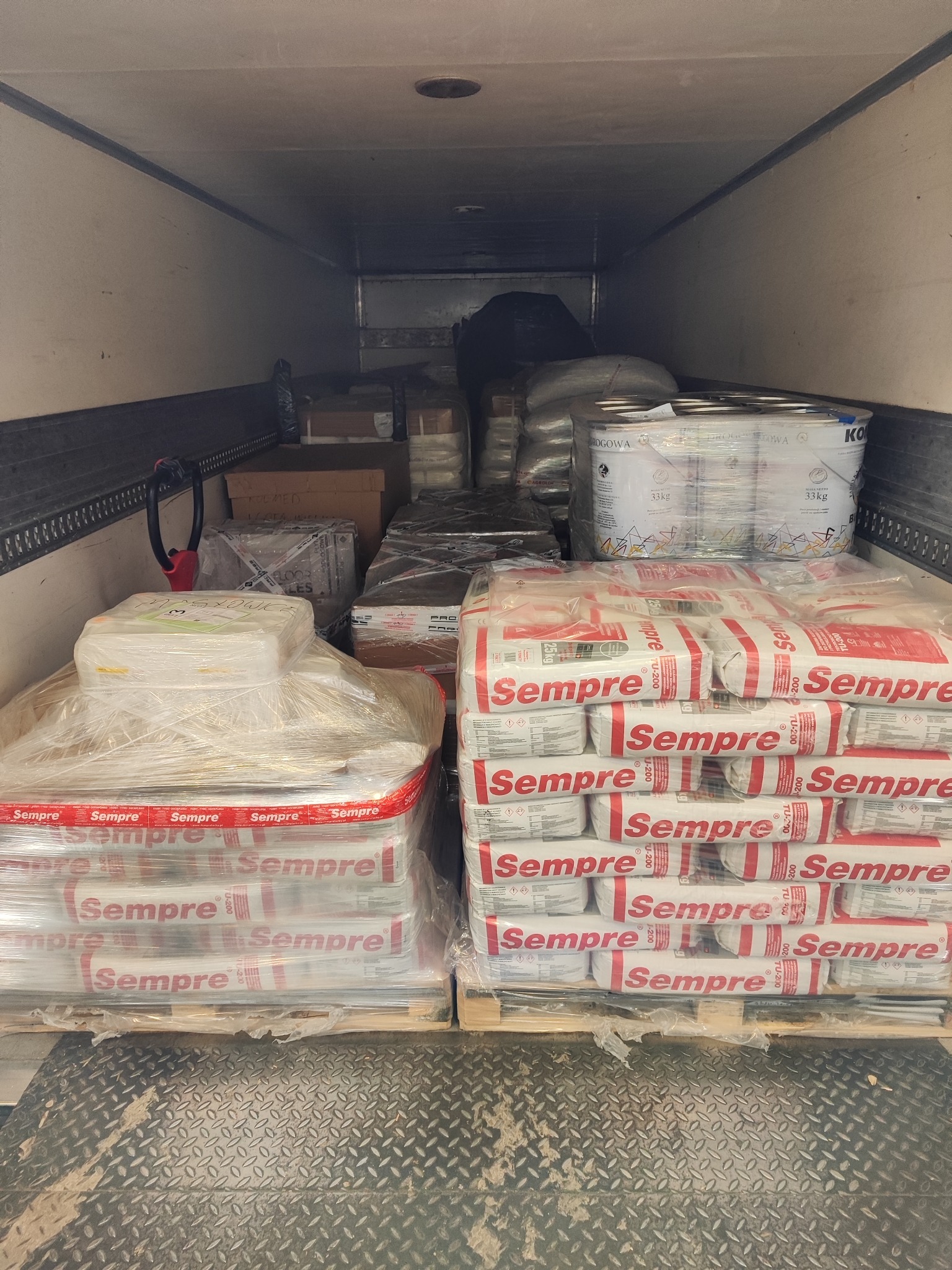 Wnętrze naczepy wypełnione paletami z zaprawami budowlanymi marki Sempre, widoczne również puszki z farbą i inne materiały budowlane, zabezpieczone folią stretch.