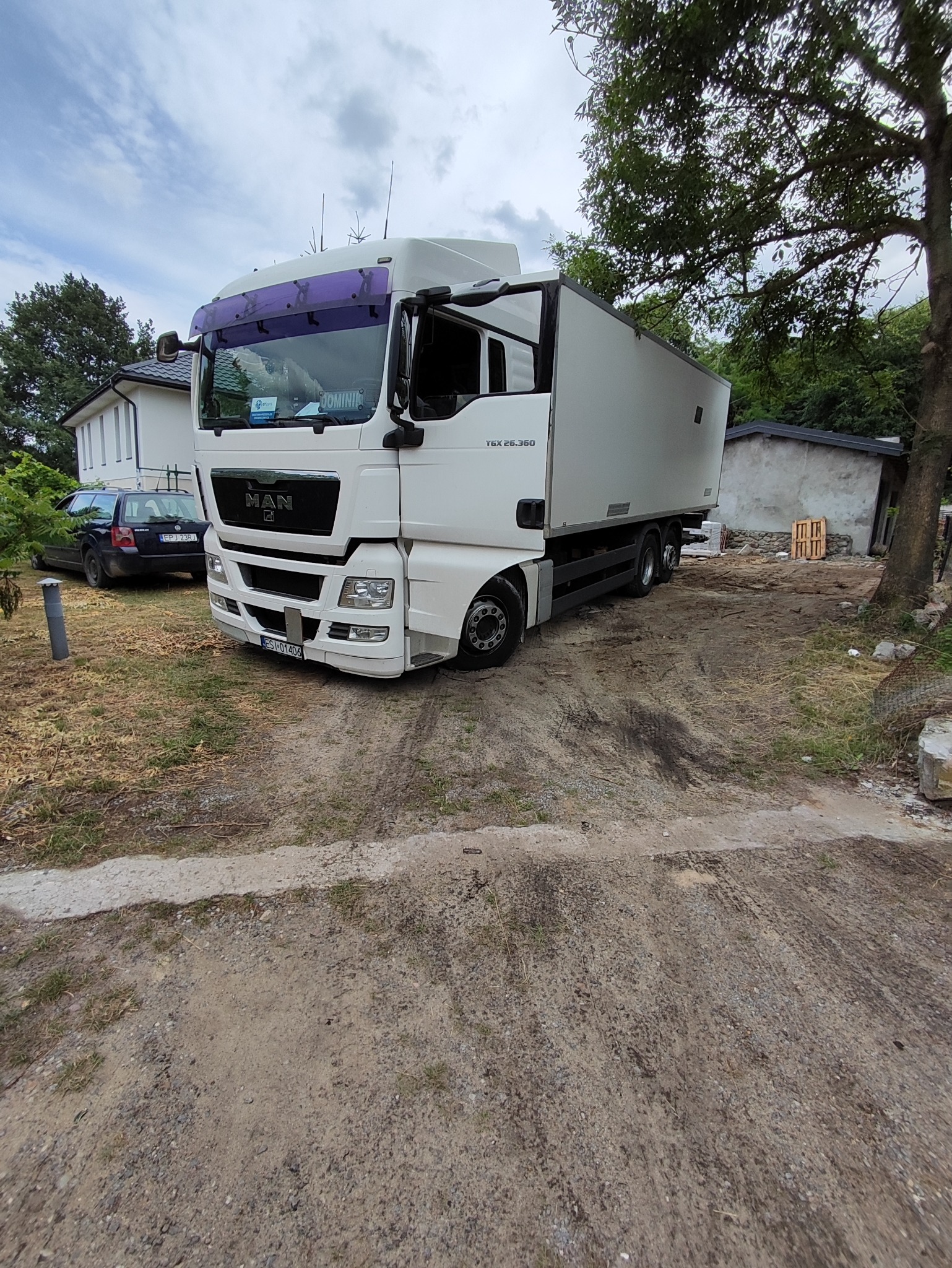 Biała ciężarówka MAN TGX 26.360 z otwartymi drzwiami kabiny, zaparkowana na żwirowym podwórku z widocznymi budynkami w tle.