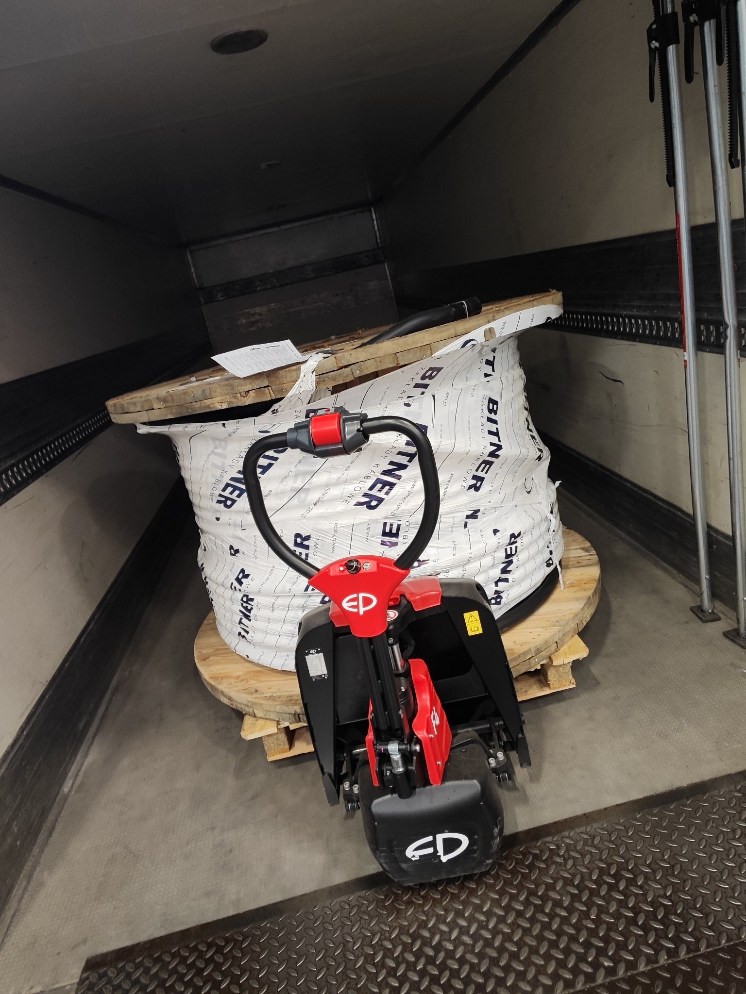 Wnętrze naczepy z paleciakiem elektrycznym przed dużą szpulą kabla elektrycznego marki Bitner, zabezpieczoną folią. Widoczne elementy mocujące ładunek.