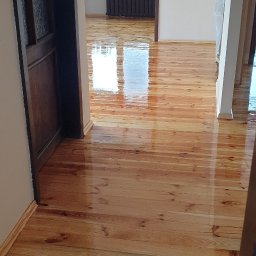 Sławomir Gniatkowski - Błyszcząca, świeżo polakierowana podłoga z jasnego drewna, widok z perspektywy korytarza, refleksy światła na powierzchni, widoczny fragment drzwi i okno w tle.