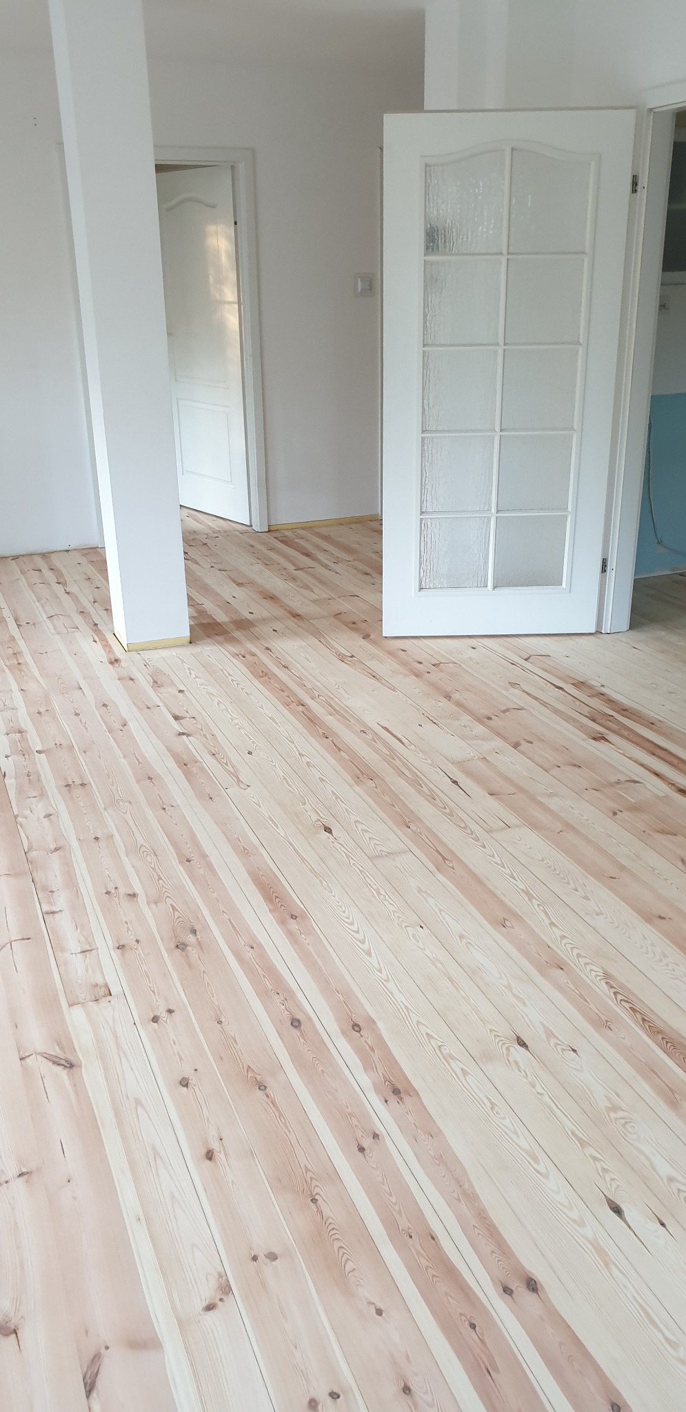 Jasne, świeżo ułożone deski podłogowe z widocznymi słojami drewna, zabezpieczone taśmą malarską przy ścianach i drzwiach.