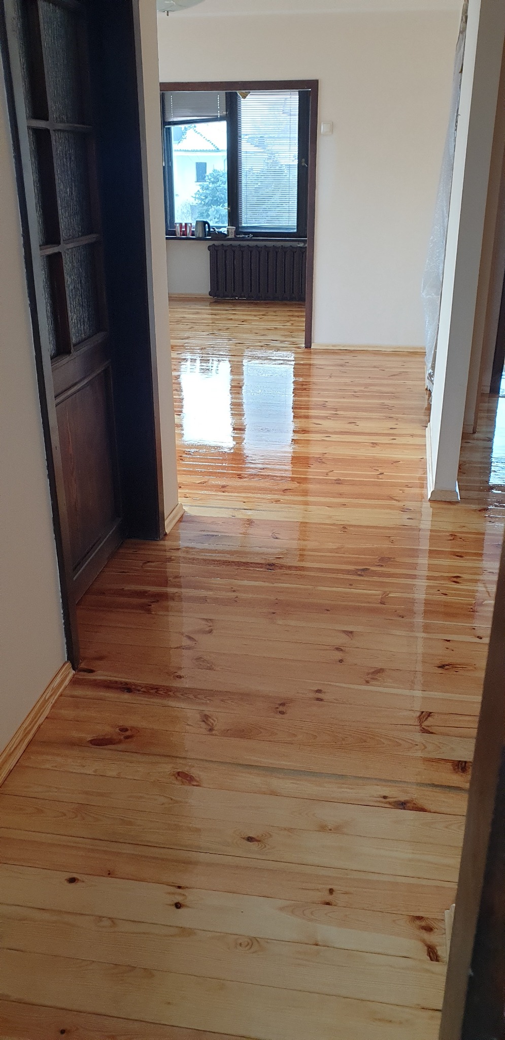 Błyszcząca, świeżo polakierowana podłoga z jasnego drewna, widok z perspektywy korytarza, refleksy światła na powierzchni, widoczny fragment drzwi i okno w tle.