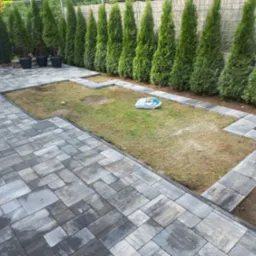 Ułożona szara kostka brukowa na posesji, obrzeża z kostki oddzielają trawnik, w tle rząd tuj przy płocie i donice. Widok z góry.