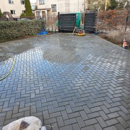 Tymoteusz Baranowski BARANOWSKI BRUK - Świeżo ułożona, mokra kostka brukowa w jodełkę na podjeździe, z widocznymi narzędziami i zagęszczarką. W tle brama wjazdowa i zieleń.