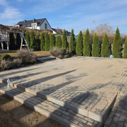 Tymoteusz Baranowski BARANOWSKI BRUK - Nowo ułożona, szara kostka brukowa na tarasie z widocznymi schodkami i butelką wody. W tle rząd tuj i plac zabaw, a dalej dom jednorodzinny pod błękitnym niebem.