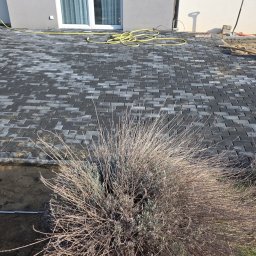 Tymoteusz Baranowski BARANOWSKI BRUK - Ułożona, szara kostka brukowa na posesji, widok z góry. W tle budynek z białymi drzwiami balkonowymi i żółty wąż ogrodowy. Na pierwszym planie uschnięte krzewy.