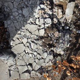 Tymoteusz Baranowski BARANOWSKI BRUK - Fragment zniszczonej betonowej nawierzchni z widocznymi pęknięciami i gruzem, częściowo usuniętej, z butem pracownika w rogu kadru i czerwonym kablem w tle.