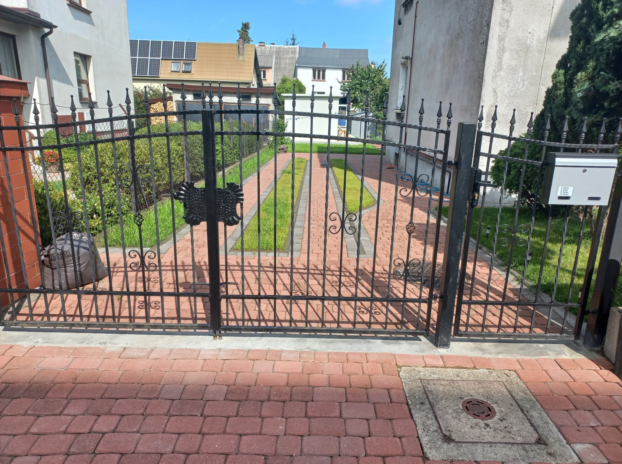 Kute, czarne ogrodzenie z otwartą bramą, widok na posesję z kostką i trawnikiem. Czerwona kostka brukowa na pierwszym planie, domy w tle, słoneczny dzień.
