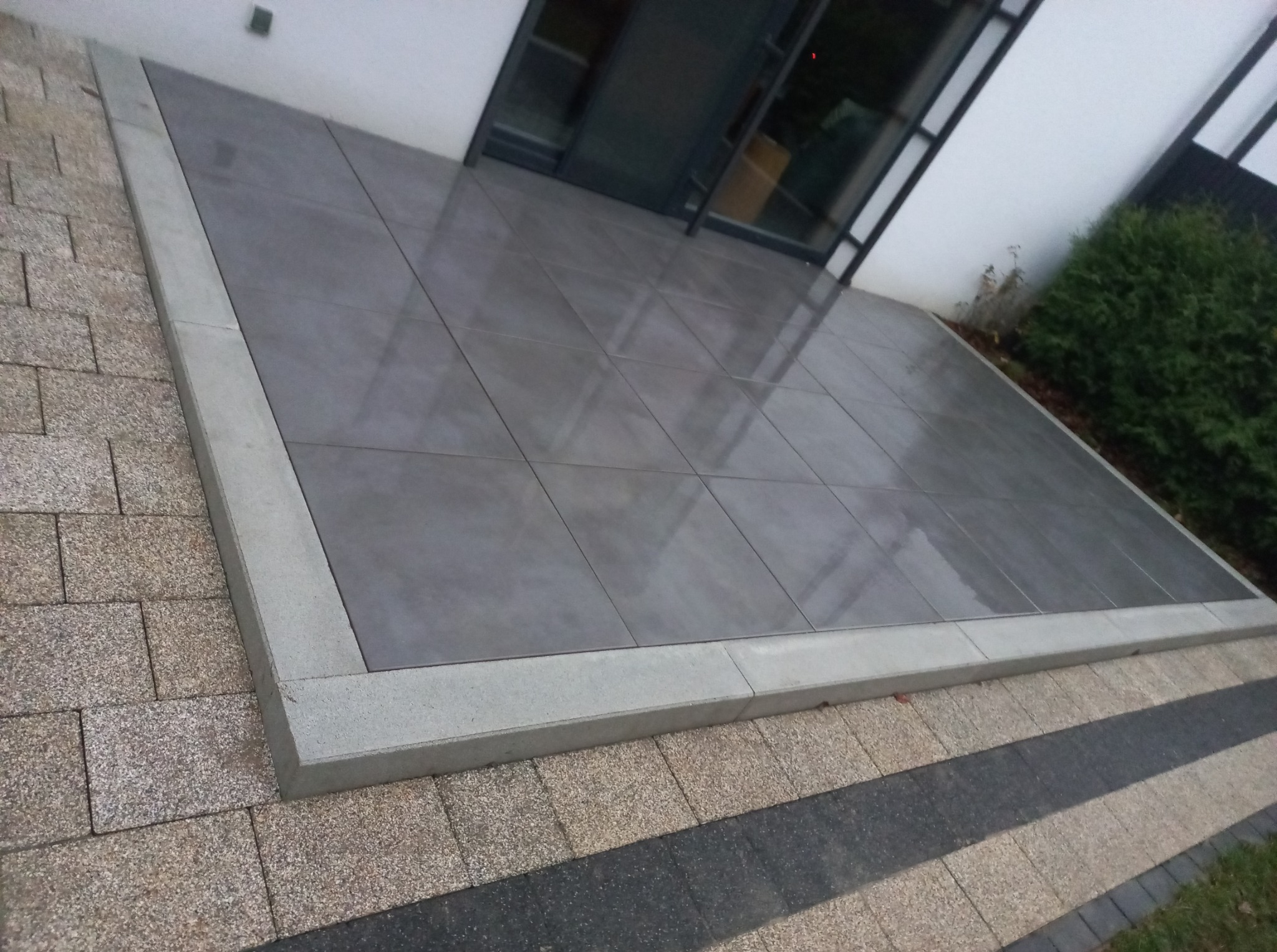 Nowoczesny taras z szarych płyt, obrzeża z jasnego betonu. Widoczne kostki brukowe w dwóch odcieniach. Minimalistyczny styl, płaska powierzchnia, świeżo ułożone.