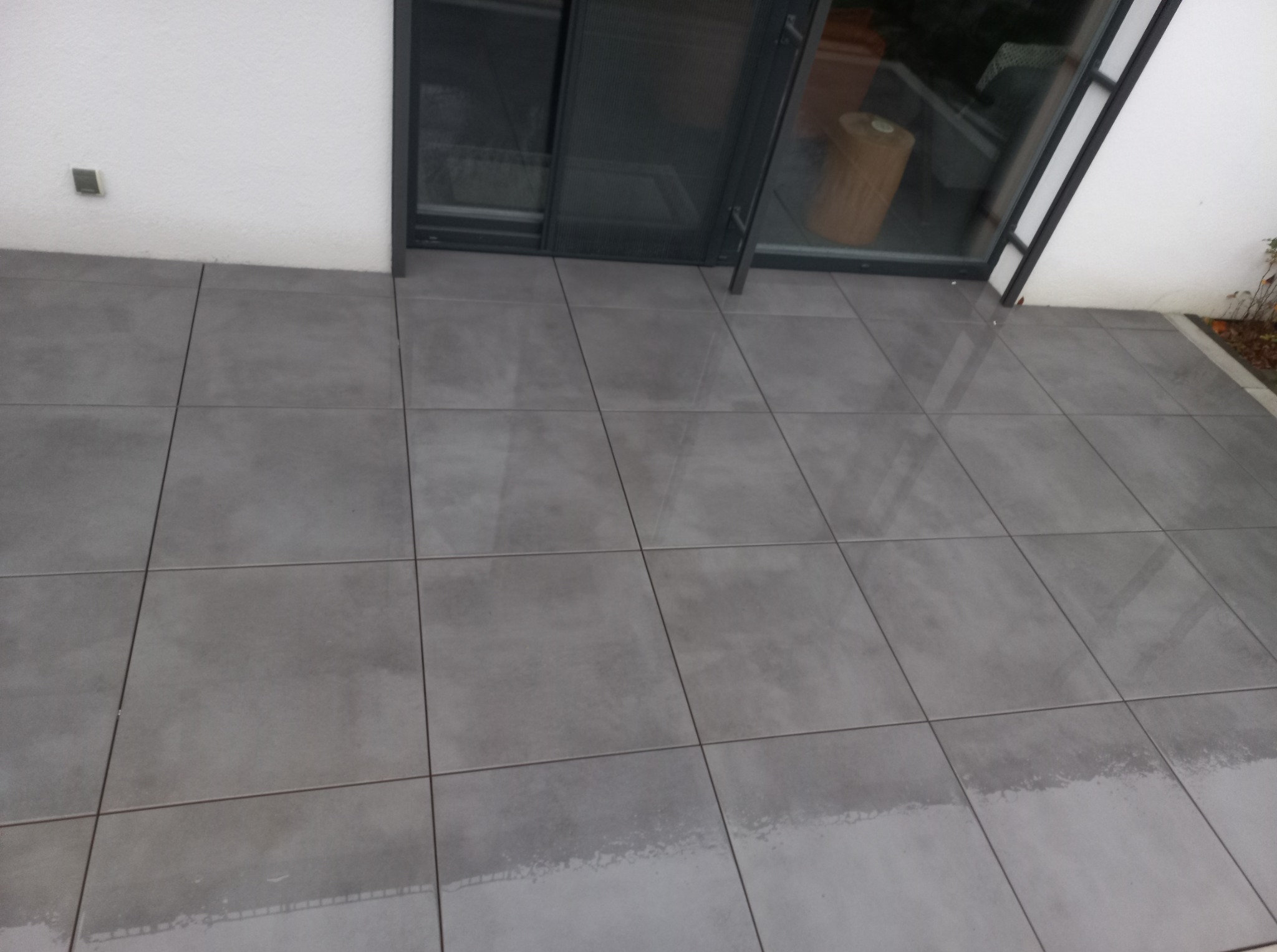 Ułożona szara, kwadratowa kostka brukowa przed wejściem do domu z przesuwnymi drzwiami. Widoczne fugi i odbicie światła na powierzchni kostki.