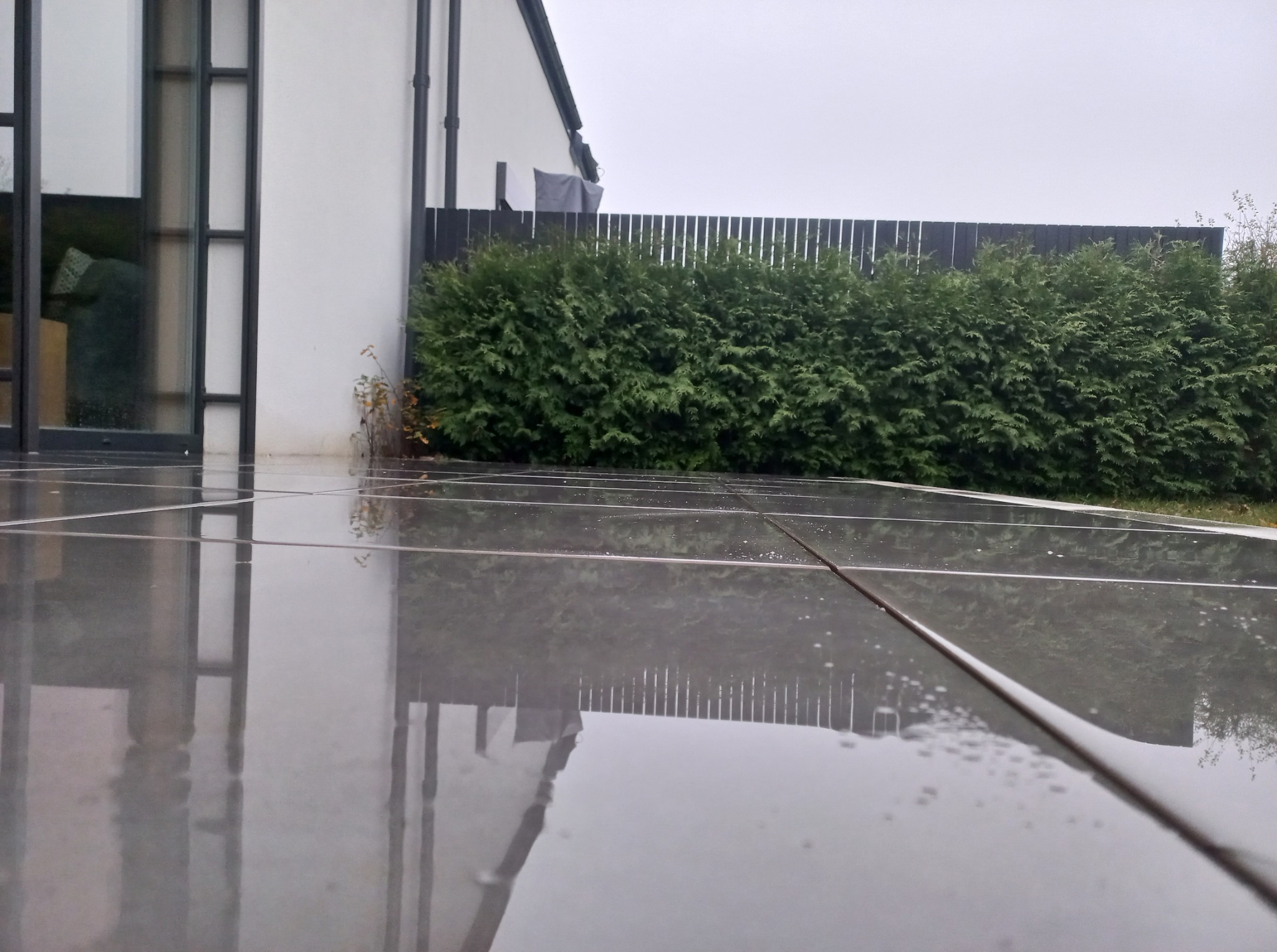 Śliska, mokra nawierzchnia z płytek na tarasie, odbijająca budynek i zieleń. Widoczna czarna, drewniana palisada i tuje. Pochmurny dzień.