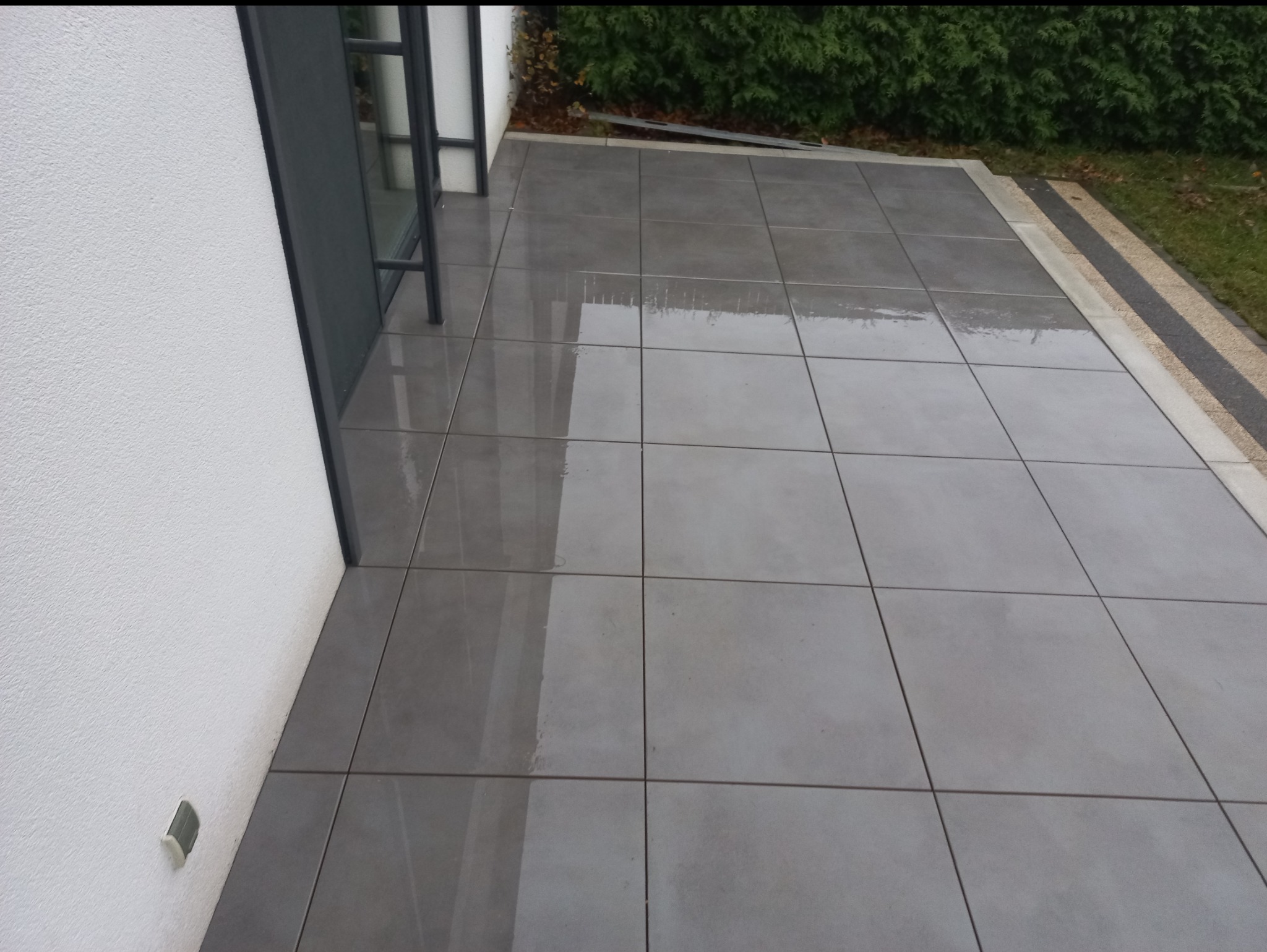 Szare, kwadratowe płyty tarasowe ułożone przy białej ścianie budynku i zielonym żywopłocie. Widoczne odbicia światła na mokrej powierzchni.