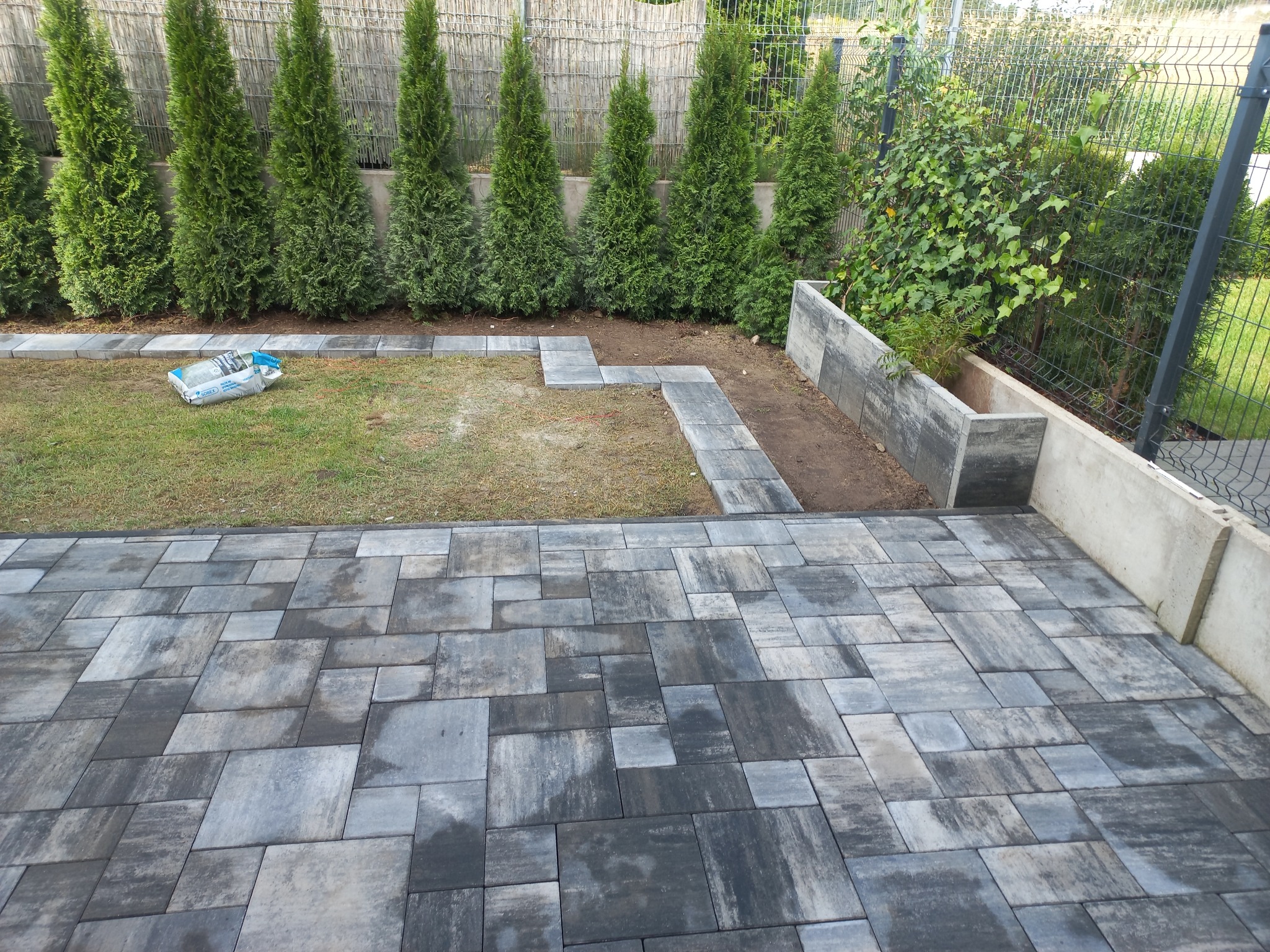 Ułożona kostka brukowa w odcieniach szarości tworzy wzór na podwórku. W tle tuje i ogrodzenie z siatki. Widoczny worek z materiałem i fragment trawnika.