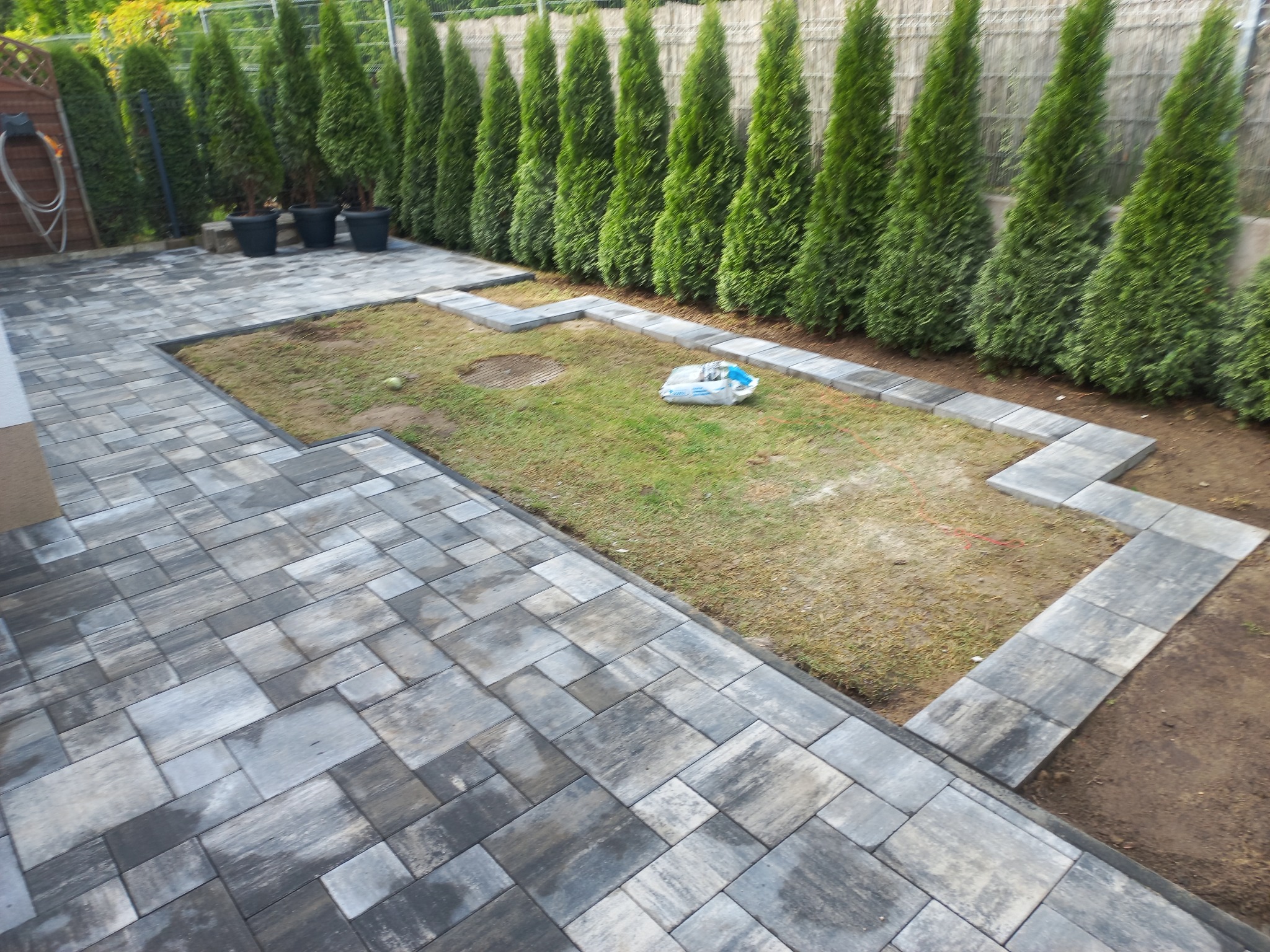 Ułożona szara kostka brukowa na posesji, obrzeża z kostki oddzielają trawnik, w tle rząd tuj przy płocie i donice. Widok z góry.