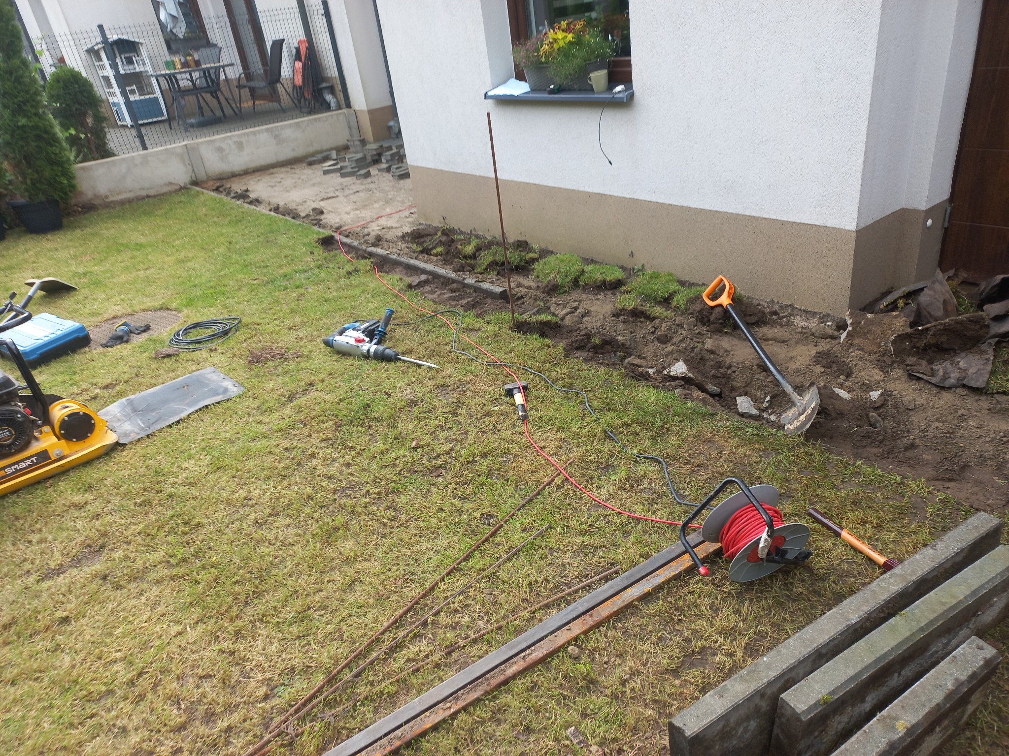 Przygotowanie podłoża pod obrzeża chodnika z kostki. Widoczny sprzęt budowlany: zagęszczarka, wiertarka, szpadel, kable i obrzeża betonowe. Częściowo ułożona kostka.