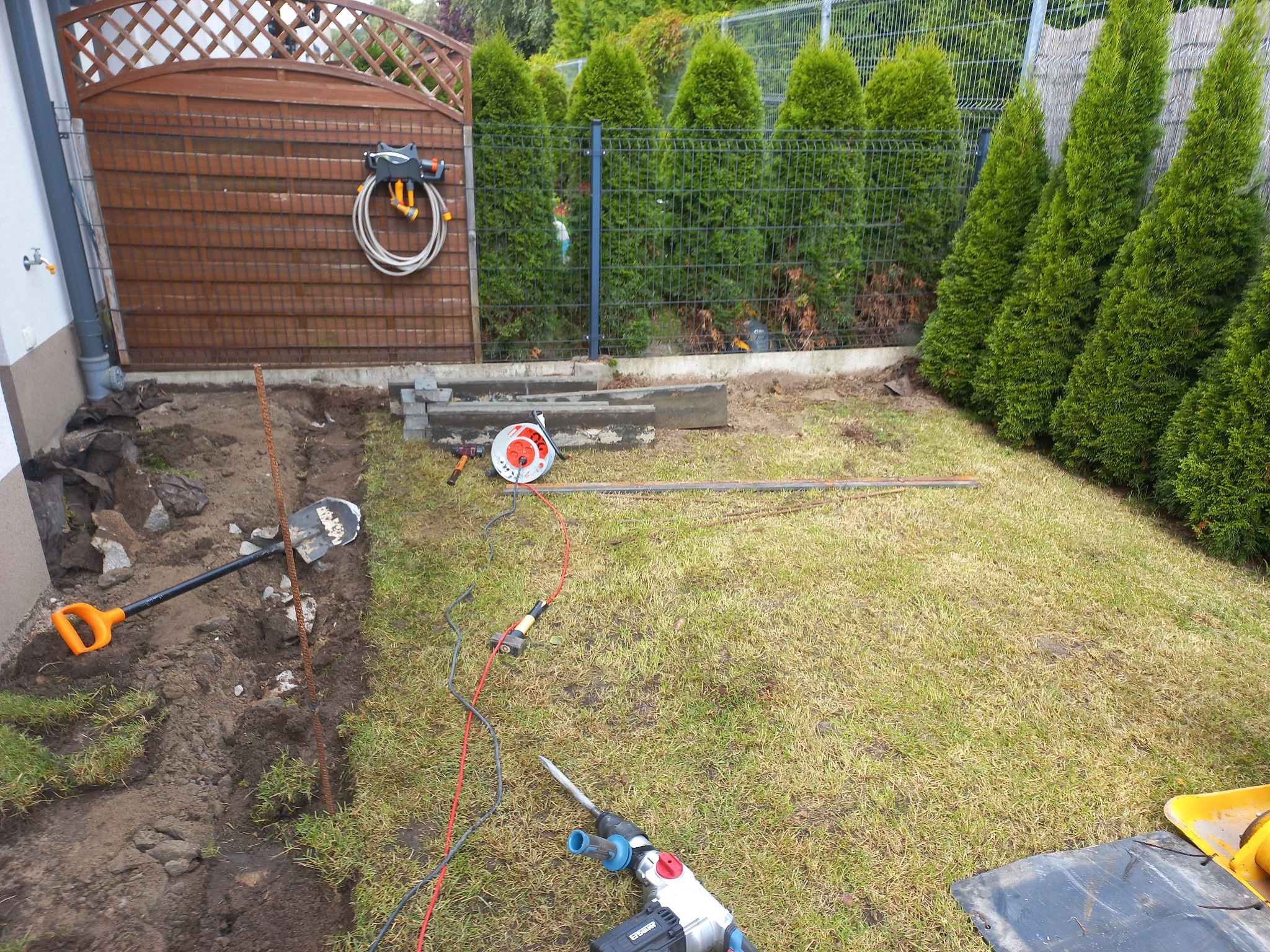 Przygotowanie terenu pod ścieżkę w ogrodzie: wykopany rów, narzędzia (szpadel, młotek, wiertarka udarowa), obrzeża kostki, trawnik, ogrodzenie z tujami w tle.