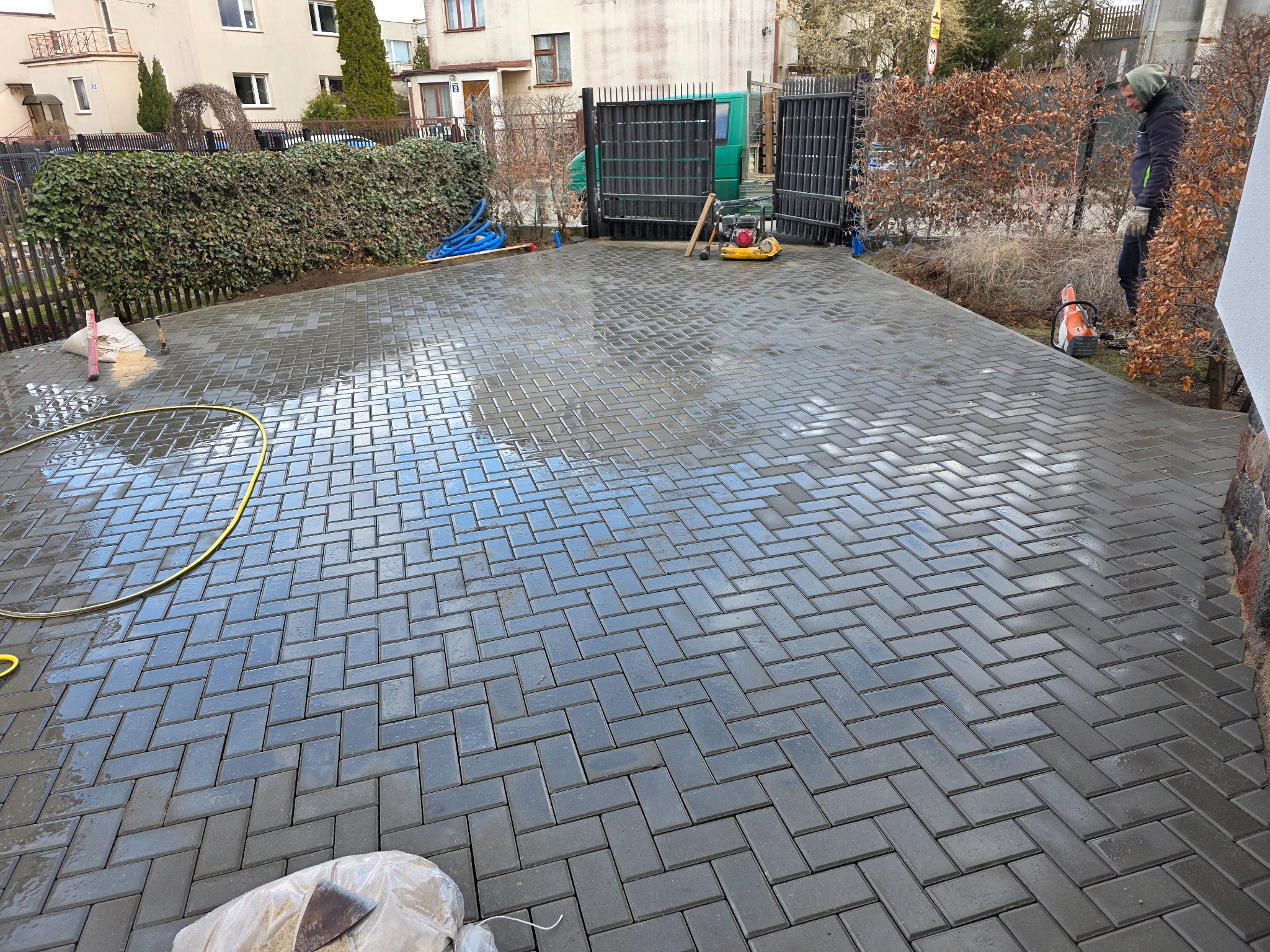 Świeżo ułożona, mokra kostka brukowa w jodełkę na podjeździe, z widocznymi narzędziami i zagęszczarką. W tle brama wjazdowa i zieleń.