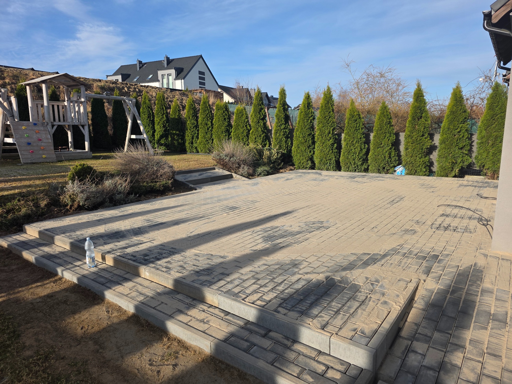 Nowo ułożona, szara kostka brukowa na tarasie z widocznymi schodkami i butelką wody. W tle rząd tuj i plac zabaw, a dalej dom jednorodzinny pod błękitnym niebem.