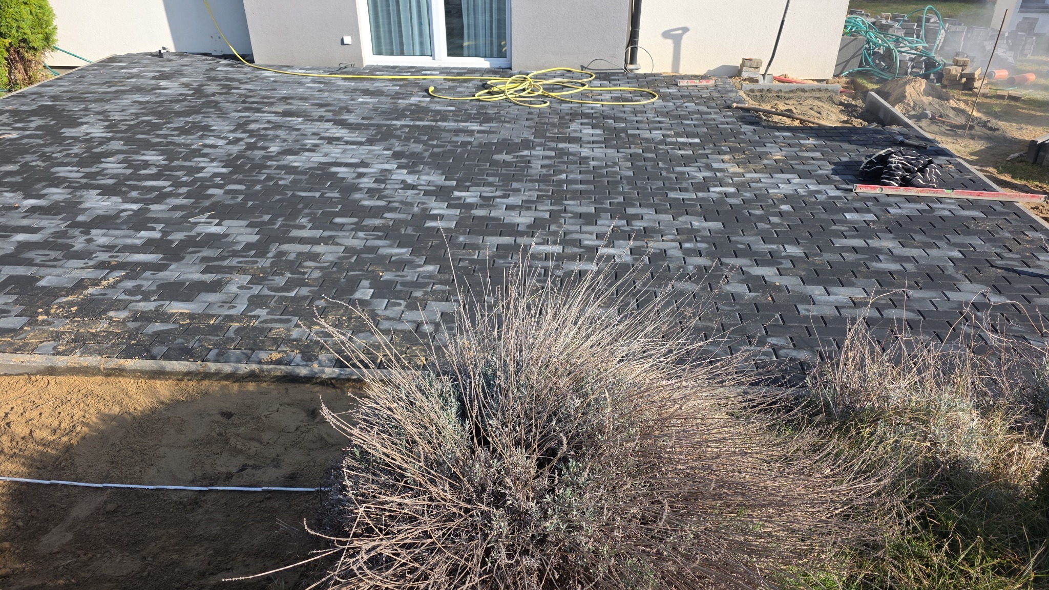 Ułożona, szara kostka brukowa na posesji, widok z góry. W tle budynek z białymi drzwiami balkonowymi i żółty wąż ogrodowy. Na pierwszym planie uschnięte krzewy.