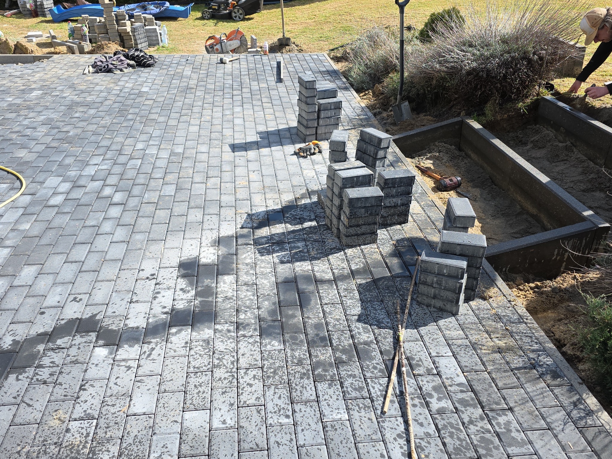 Fragment układanej szarej kostki brukowej na posesji, z widocznymi stosami kostki, narzędziami i przygotowanym obrzeżem rabaty. Częściowo ułożona nawierzchnia.