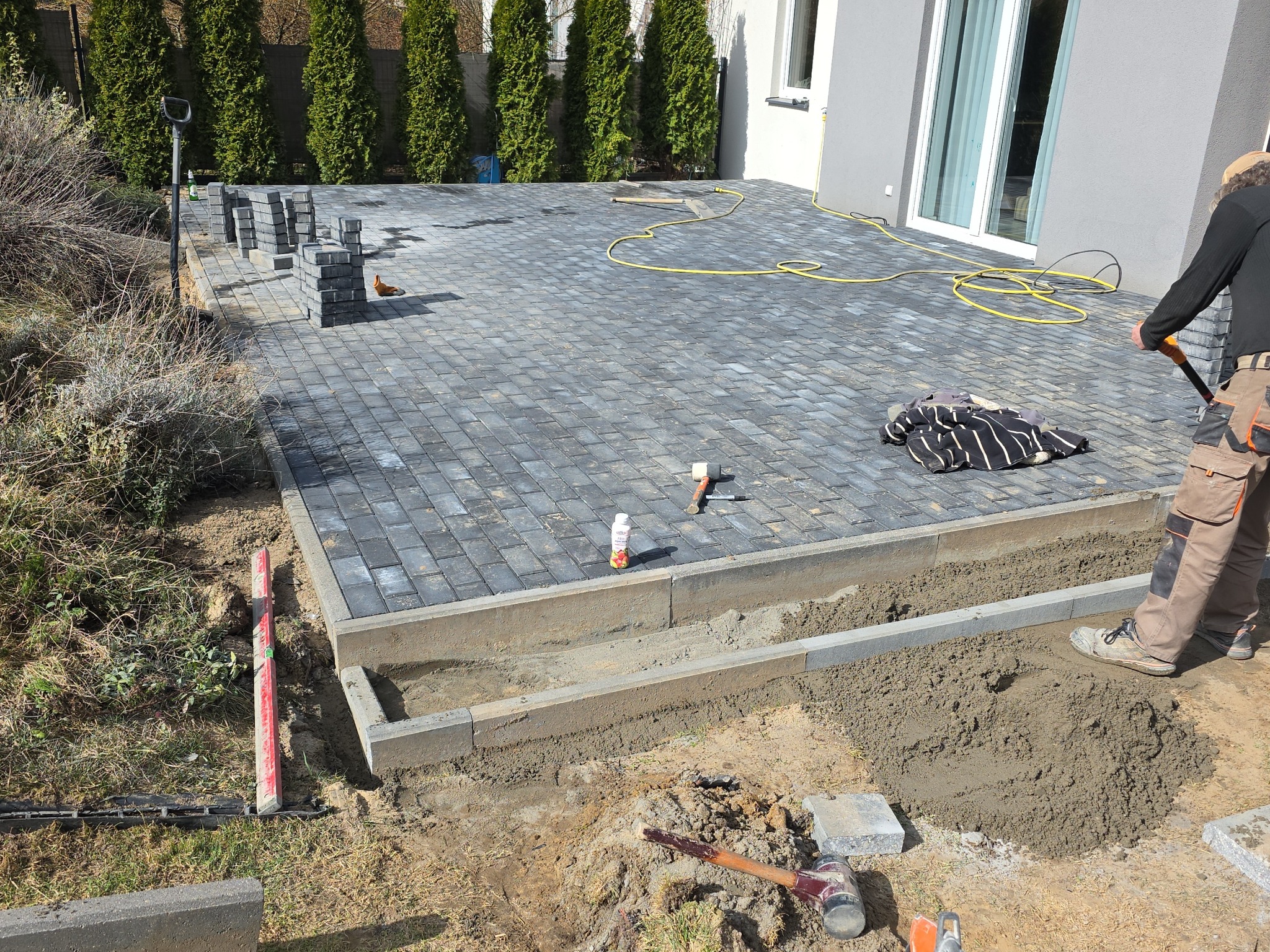 Fragment układania kostki brukowej na posesji. Widoczne ułożone kostki, narzędzia pracy, oraz pracownik w trakcie wykonywania prac brukarskich.