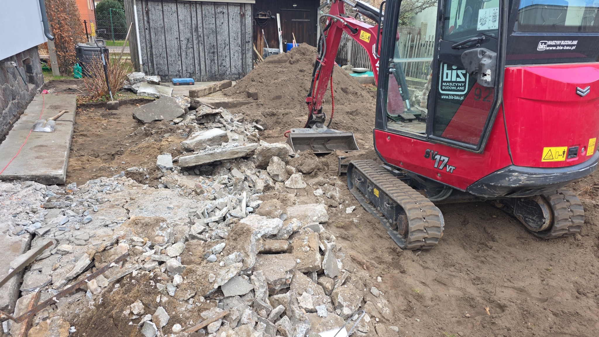 Czerwona mini koparka na placu budowy, gruz i ziemia. Widoczne fragmenty fundamentów i narzędzia. Etap prac rozbiórkowych.