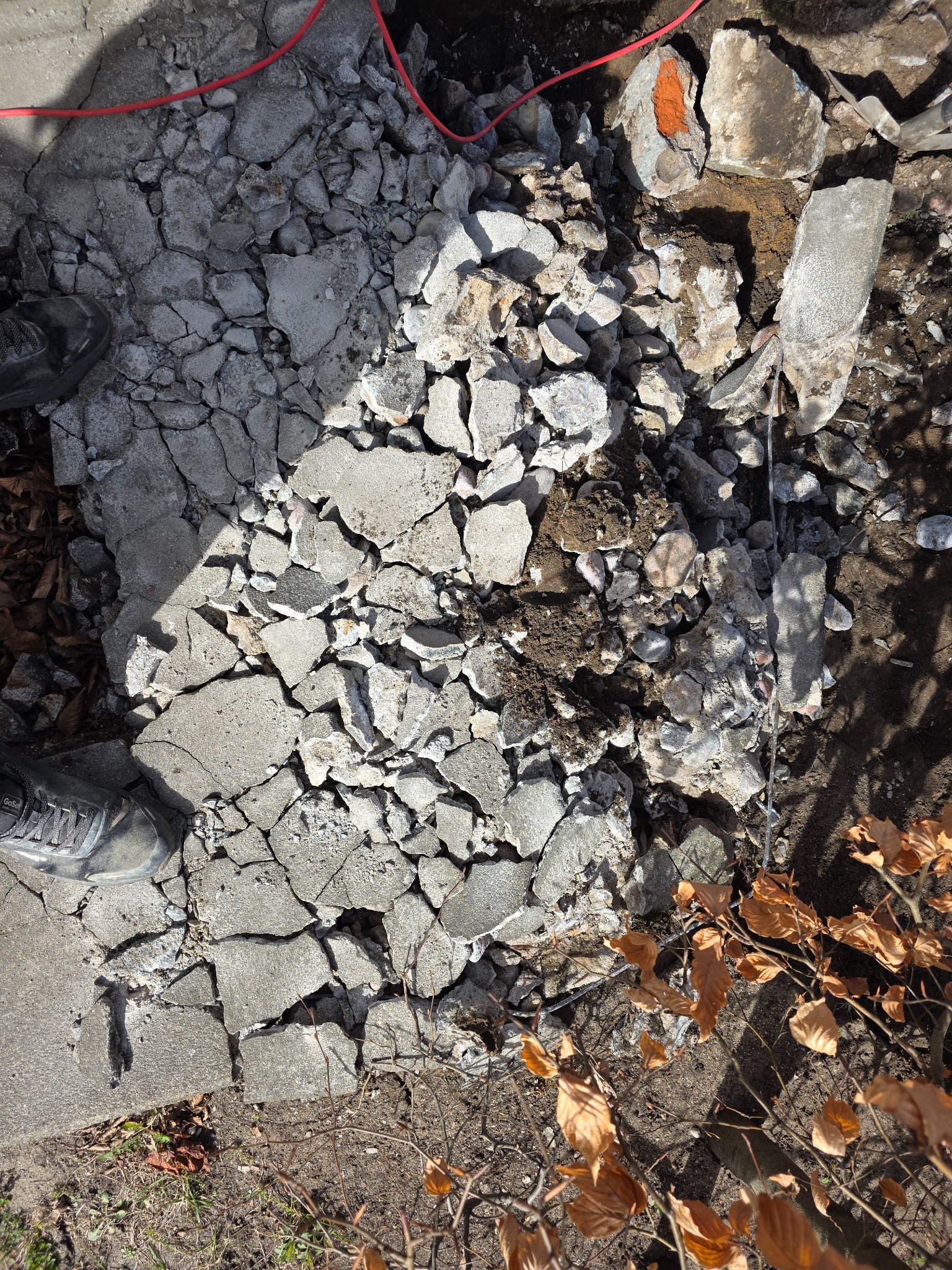 Fragment zniszczonej betonowej nawierzchni z widocznymi pęknięciami i gruzem, częściowo usuniętej, z butem pracownika w rogu kadru i czerwonym kablem w tle.