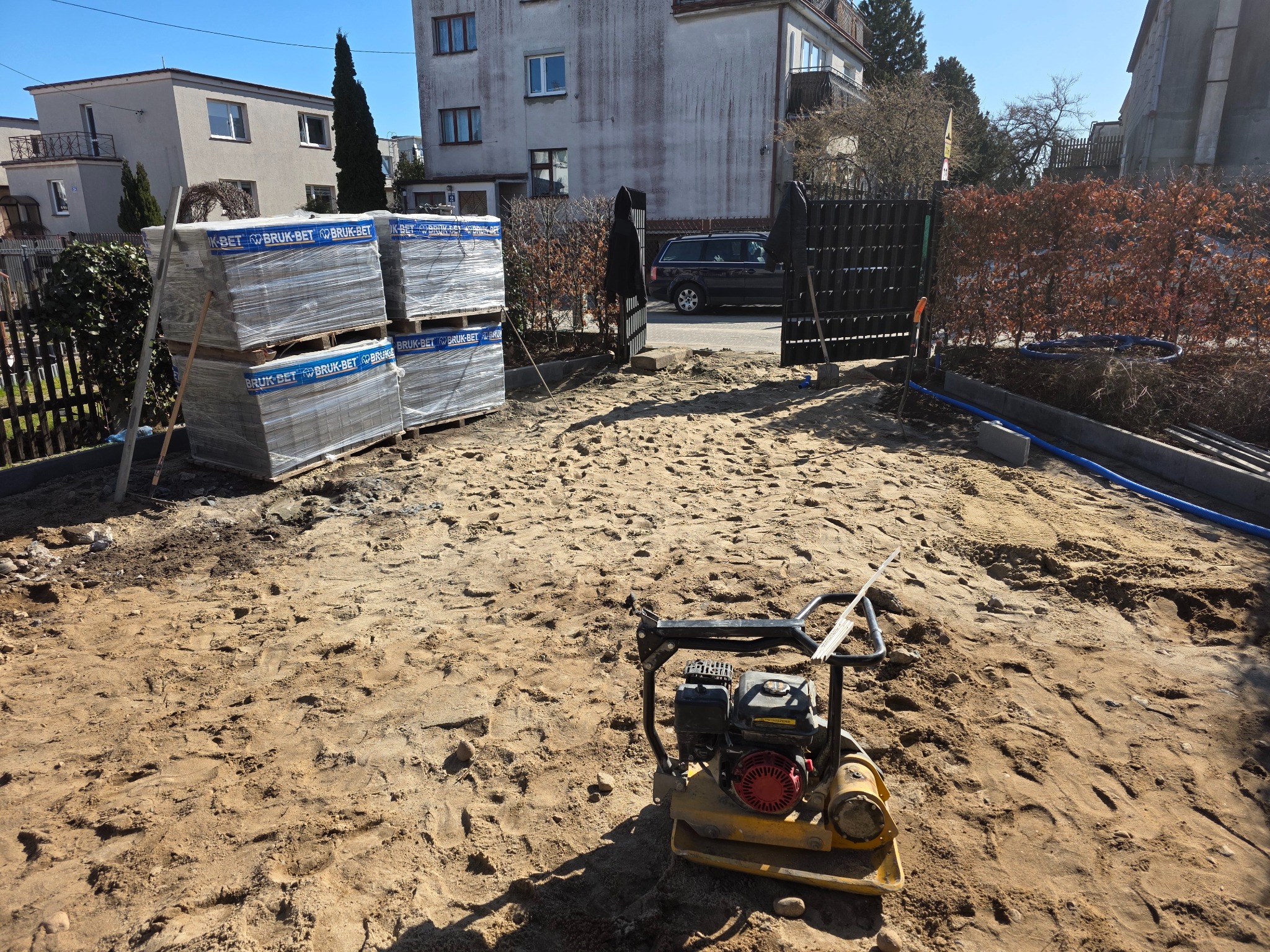 Przygotowanie terenu pod kostkę: ubijarka, palety z kostką Bruk-Bet, piasek. Widok na posesję z bramą i budynkami w tle, słoneczny dzień.