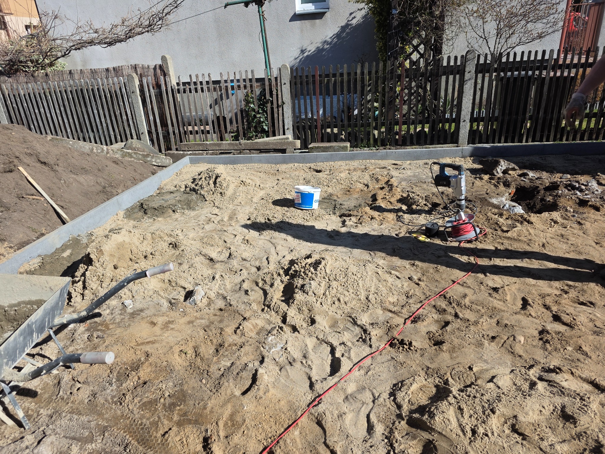 Przygotowanie terenu pod kostkę brukową w Bolszewie. Widoczny piasek, obrzeża, narzędzia i częściowo taczka. Prace ziemne w toku, ogrodzenie w tle.