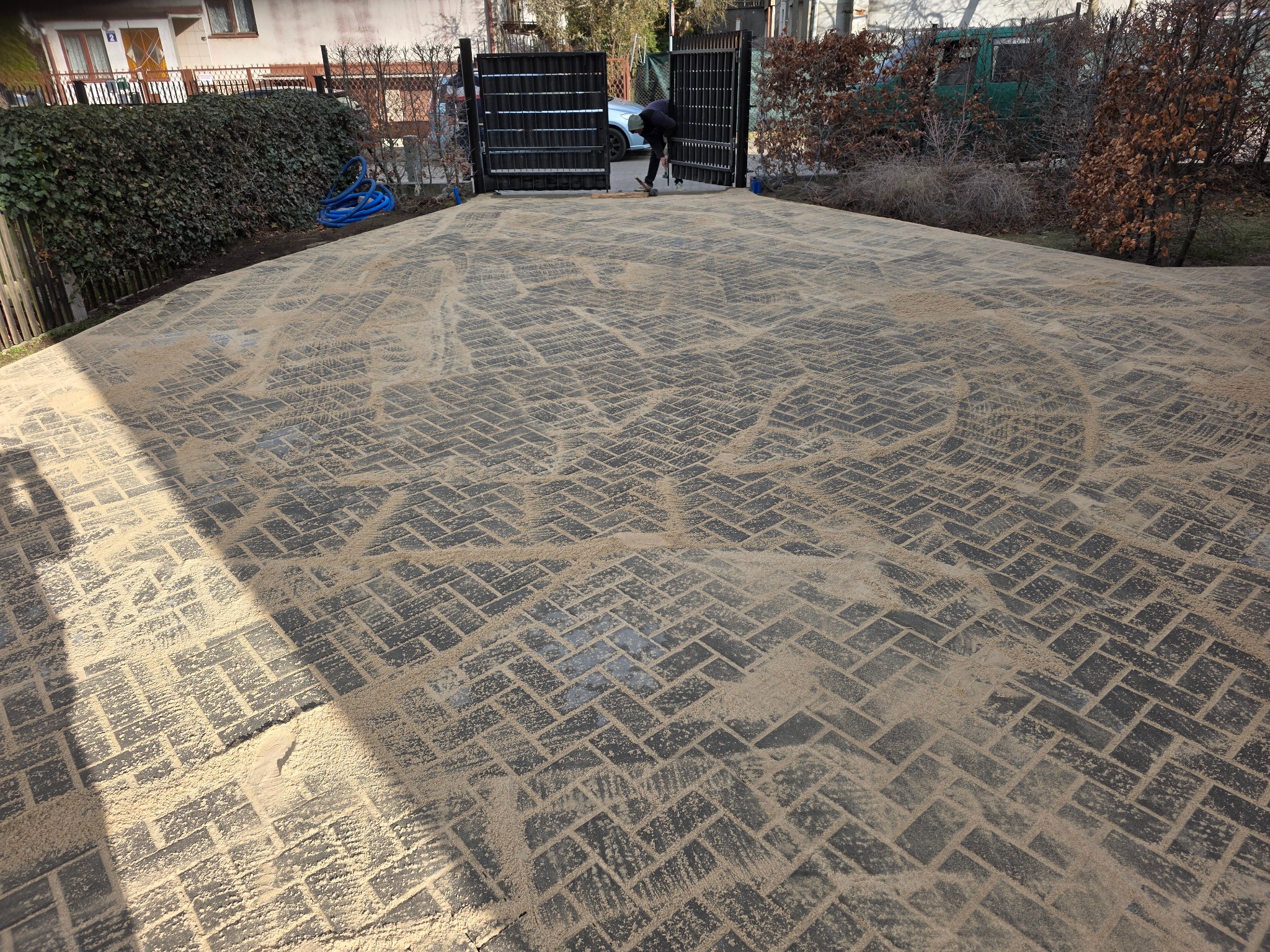 Ułożona kostka brukowa w jodełkę, zasypana piaskiem, z widoczną furtką i częściowo sylwetką osoby podczas prac wykończeniowych. Całość w słoneczny dzień.