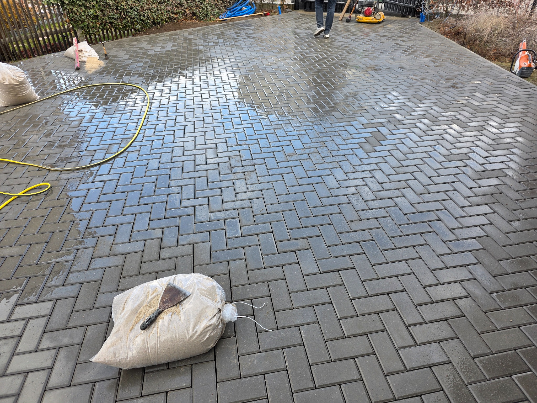Ułożona, mokra kostka brukowa w jodełkę, z workiem piasku i narzędziem. W tle osoba obsługująca zagęszczarkę, co sugeruje trwający proces układania.