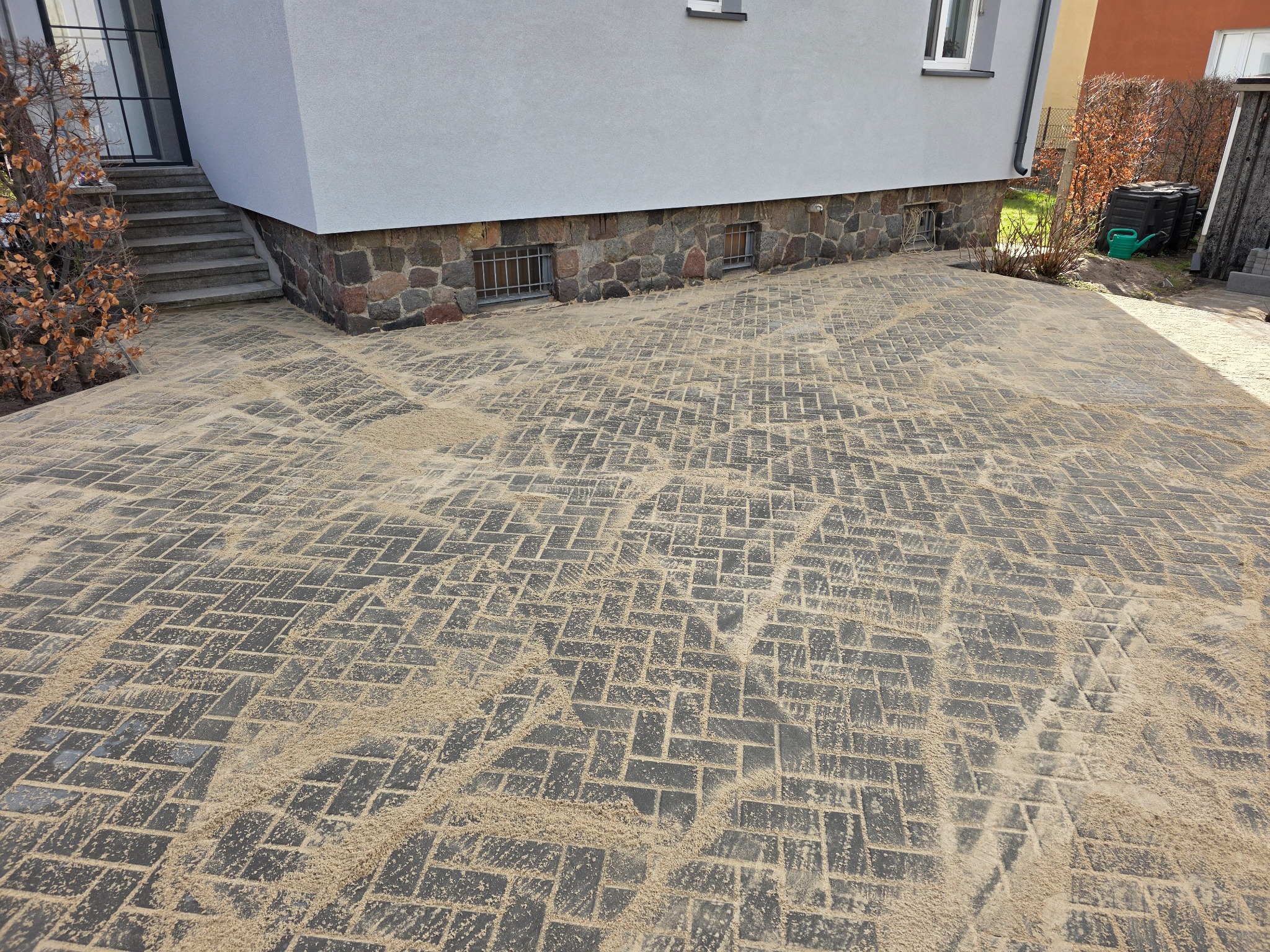 Świeżo ułożona, szara kostka brukowa w jodełkę, zasypana piaskiem, przylegająca do budynku z kamienną podmurówką i oknami z kratami. Widok z góry.