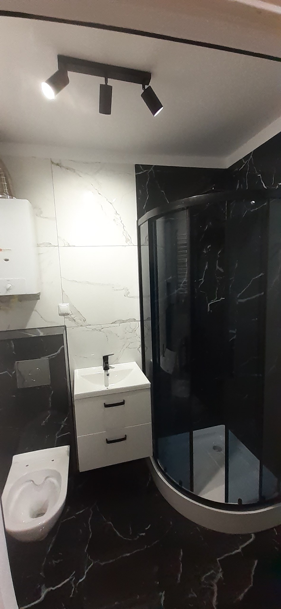 Wykończona łazienka z czarno-białymi marmurkowymi płytkami, kabiną prysznicową z czarnymi ramami, białą szafką umywalkową z czarnymi uchwytami i podwieszaną toaletą, oświetlona trzypunktowym...