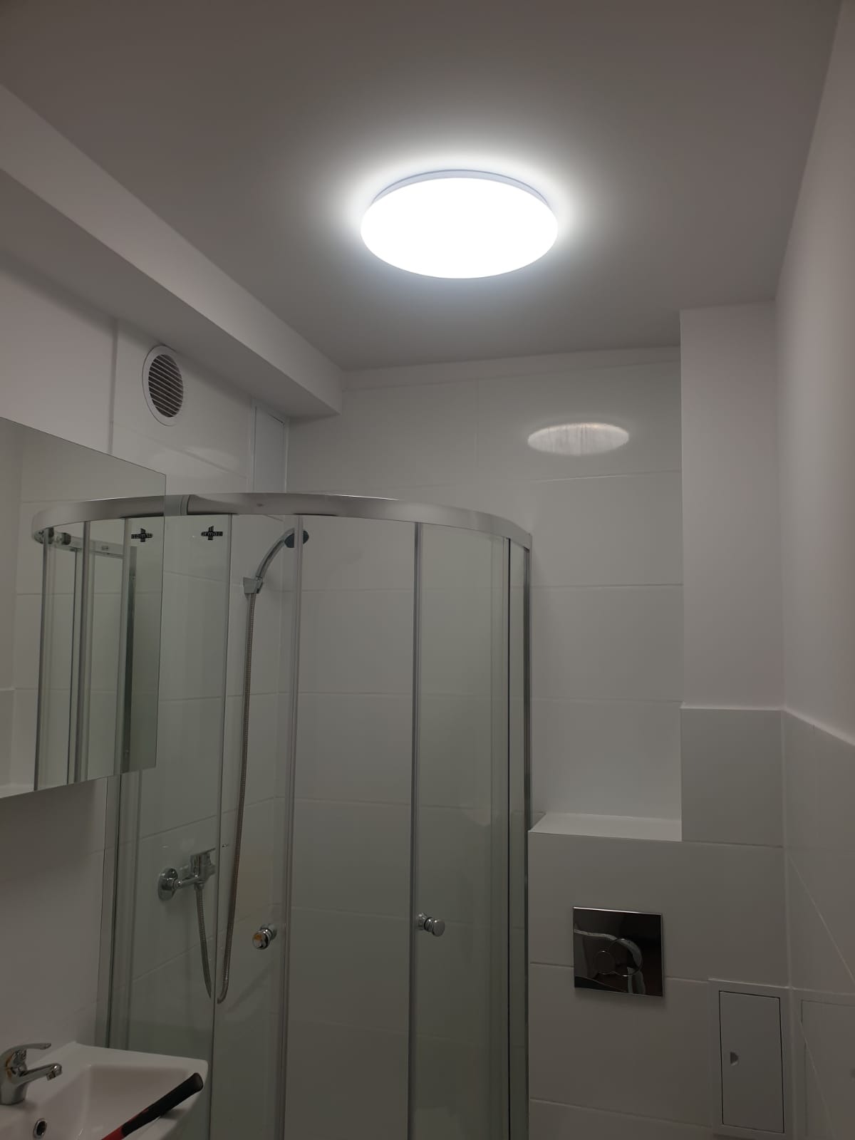Biała łazienka po remoncie z kabiną prysznicową z zaokrąglonymi drzwiami, widoczną armaturą, umywalką oraz okrągłym oświetleniem sufitowym LED.
