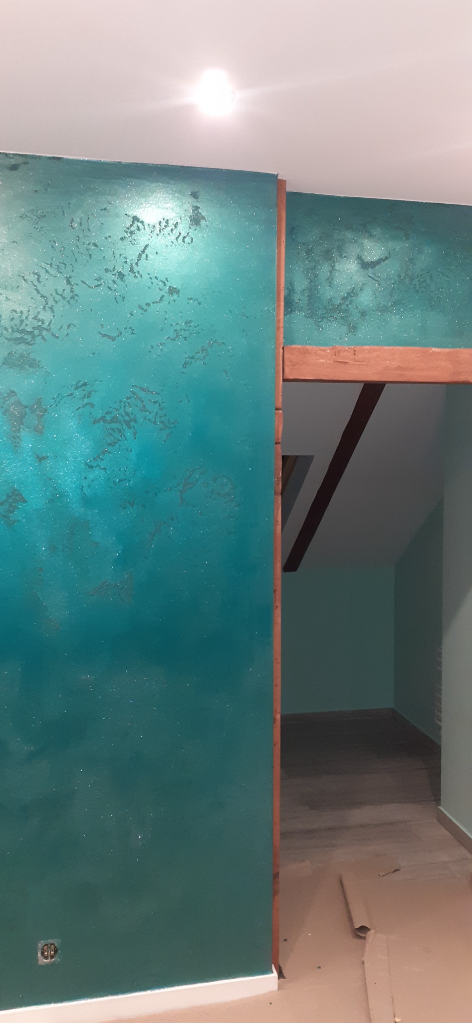 Ściana w kolorze turkusowym z efektem dekoracyjnym, widoczna rama drewniana drzwi i fragment podłogi zabezpieczonej kartonem.