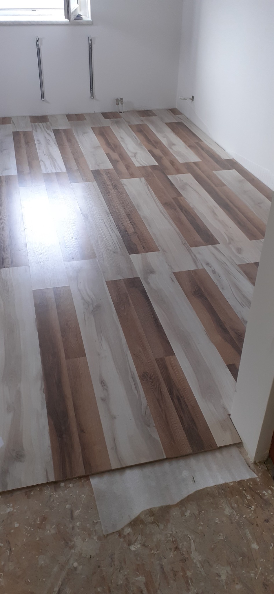 Układanie paneli podłogowych w odcieniach brązu i beżu, widoczny fragment podłogi przed położeniem paneli z resztkami kleju i zabrudzeń.